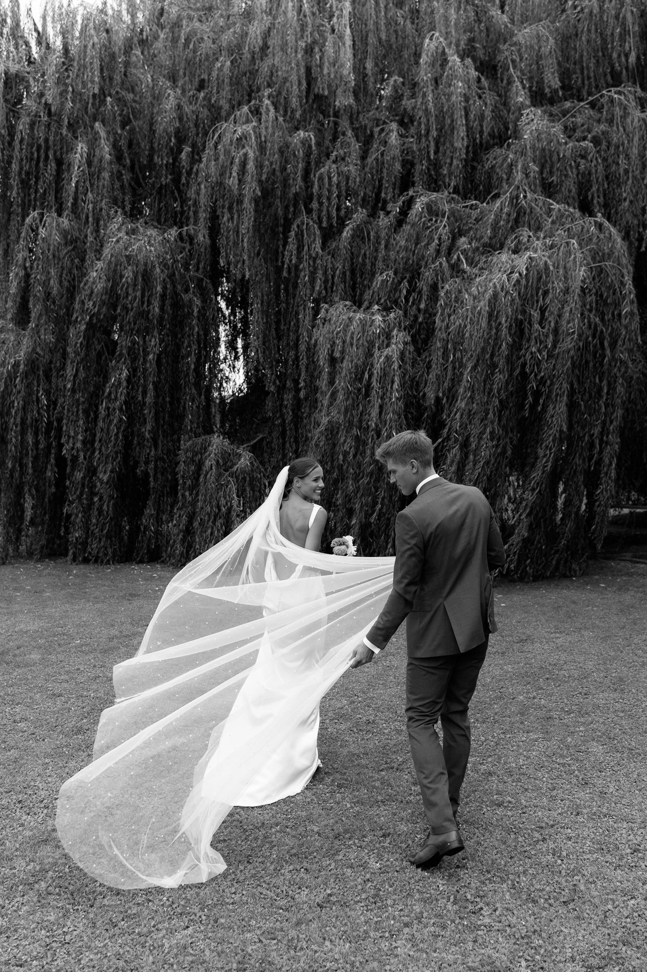 Black and white photo of bride and groom walking under weeping willow, veil flowing gracefully in the breeze.