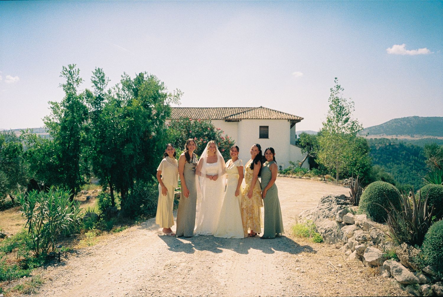 Bride and bridesmaids in gold dresses posing together outdoors at a rustic wedding venue with greenery and hills.