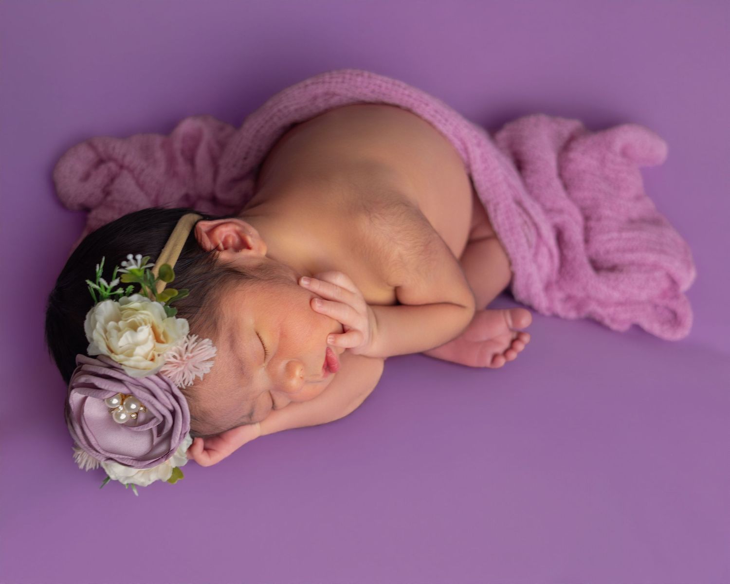 Sleeping newborn baby girl on purple background wearing a floral headband, wrapped in a soft pink knit blanket.