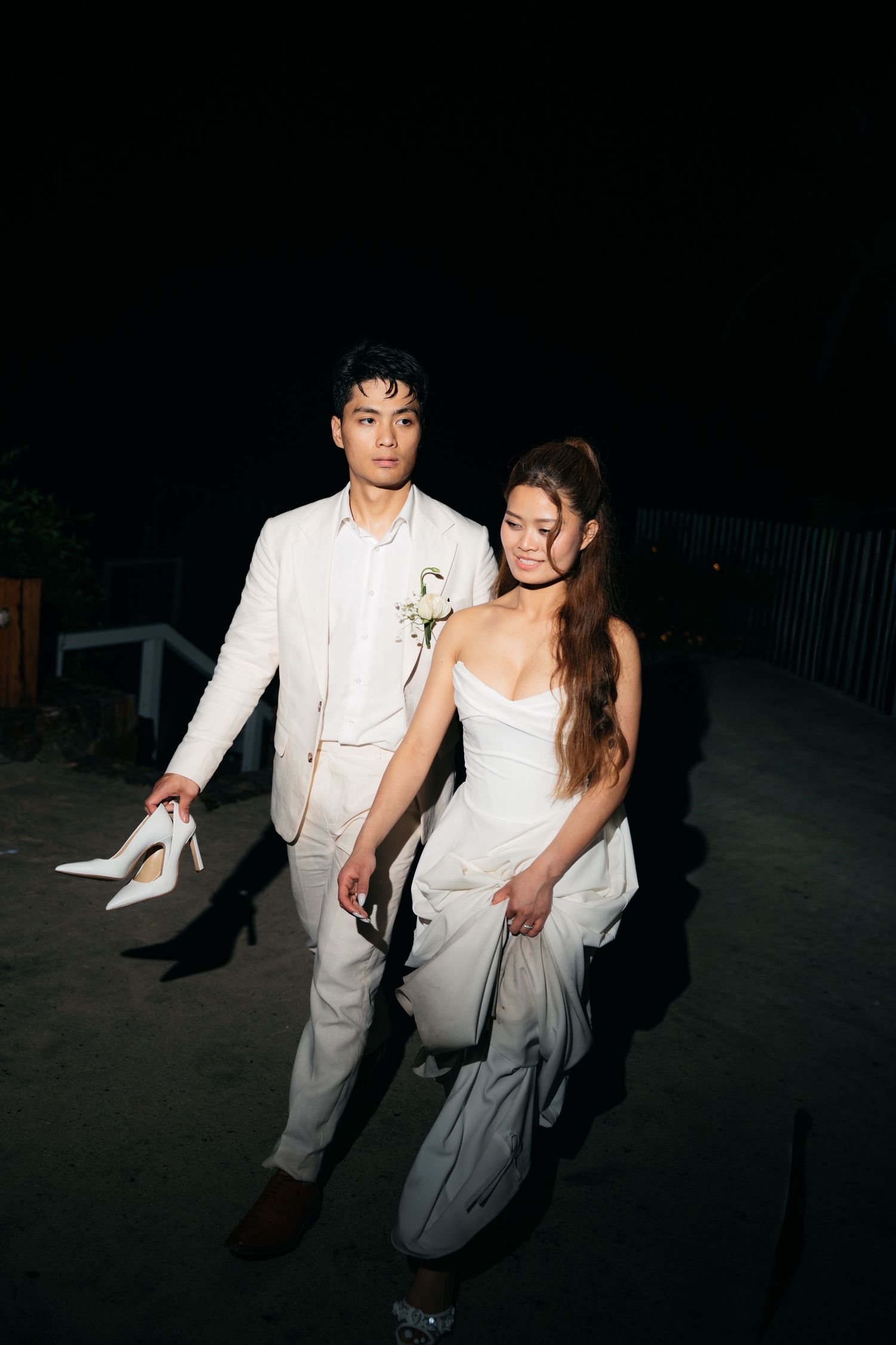 A couple in white wedding attire walks at night, the groom carrying heels and wearing a floral boutonniere.