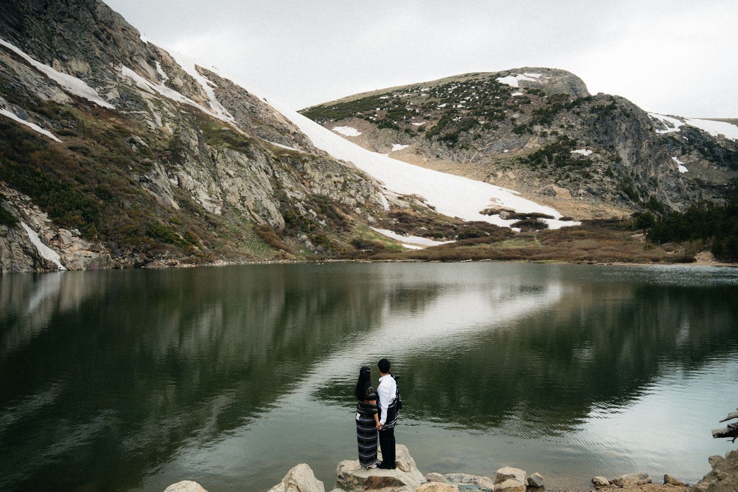 Couple standing on rocks beside a serene alpine lake surrounded by snow-dusted mountains under overcast skies.