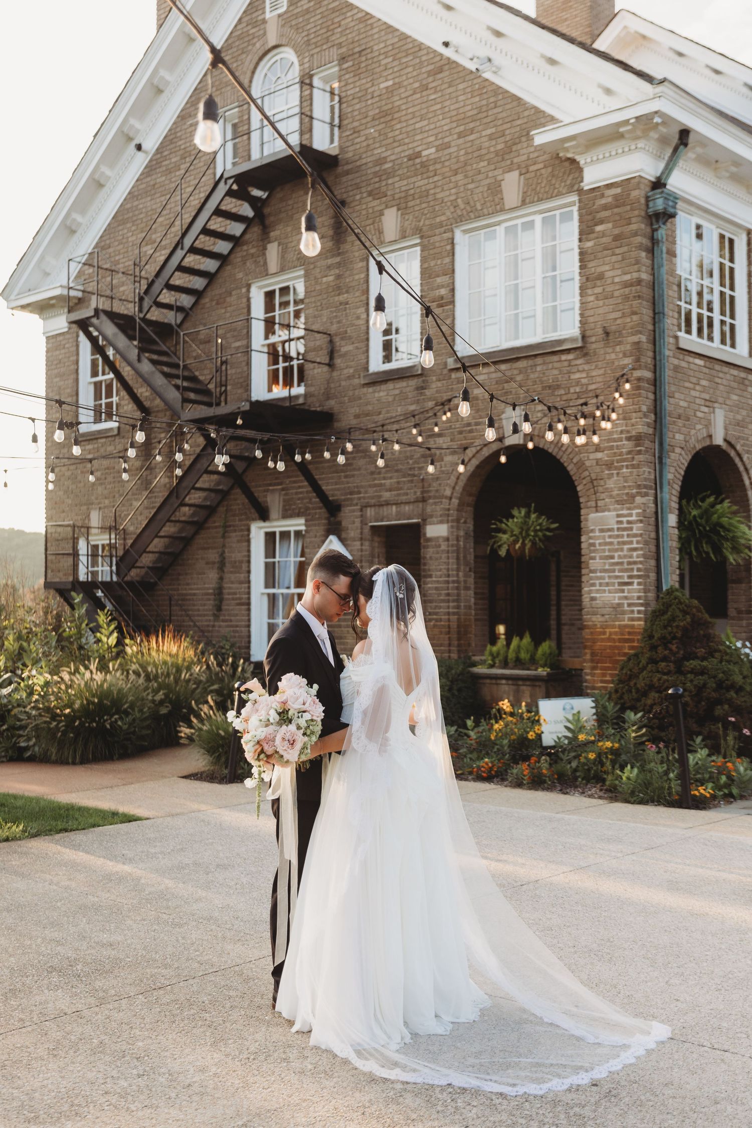 Bride and groom share romantic moment outside elegant brick venue with string lights and fire escape staircase.