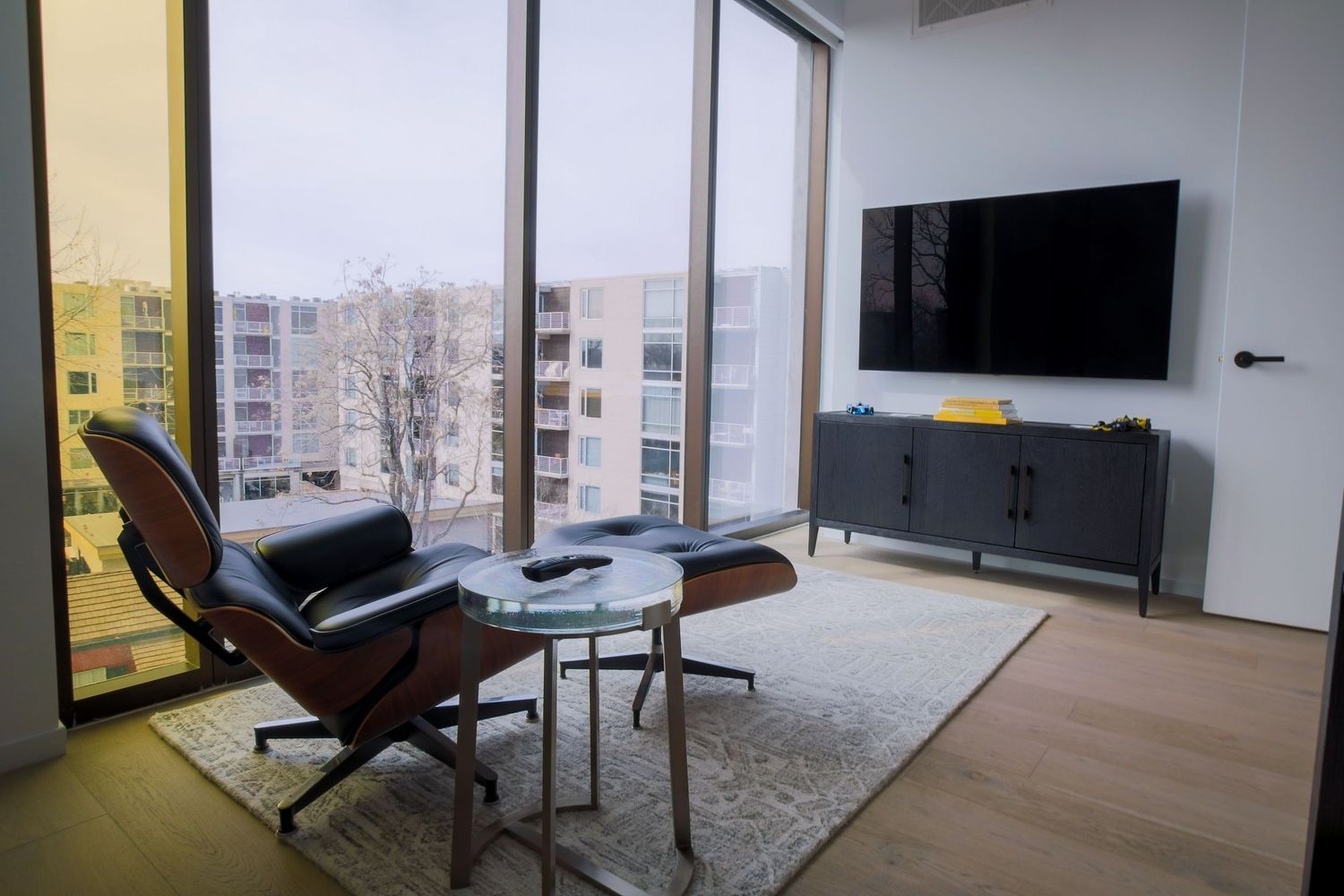 Modern living room with Eames lounge chair, flat-screen TV, and floor-to-ceiling windows overlooking city buildings.