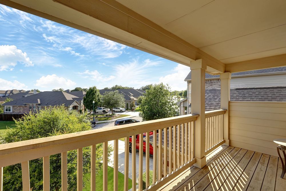 Wooden deck balcony with railing overlooking a suburban neighborhood with green trees and blue sky.