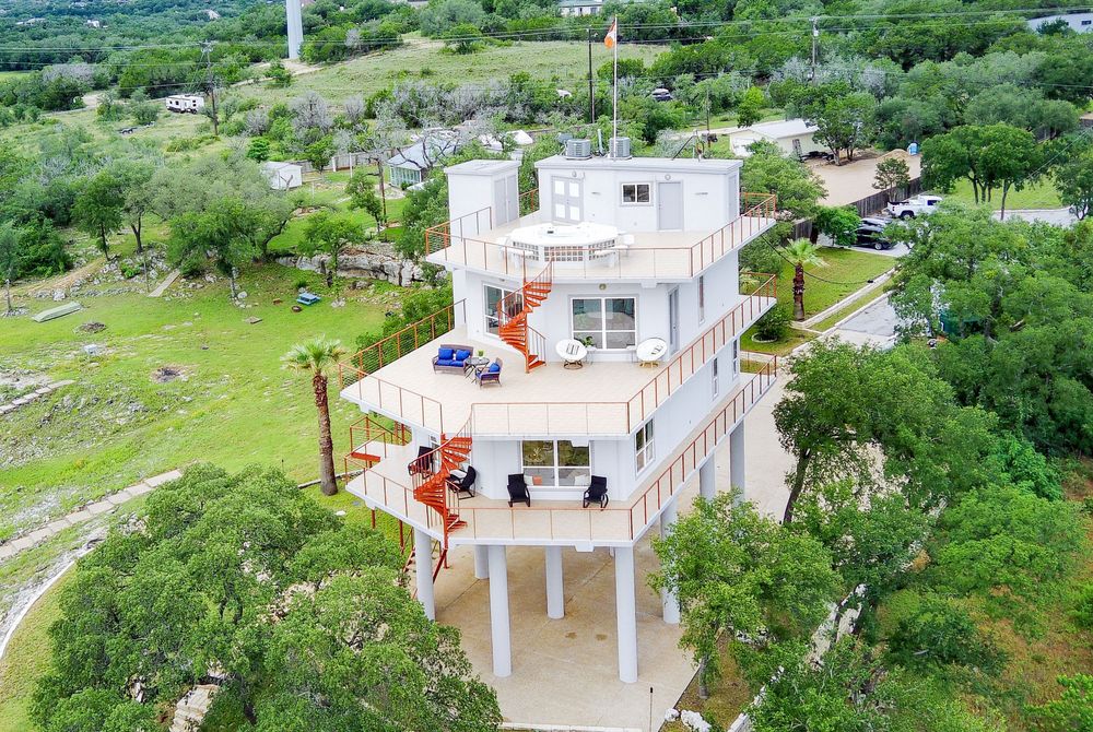 Aerial view of a multi-story white vacation home with wraparound decks and orange staircase surrounded by lush Texas Hill Country trees.