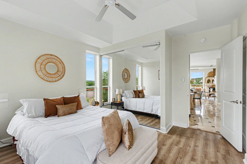 Bright bedroom with white bedding, brown accent pillows, ceiling fan, hardwood floors, and large windows with natural light.