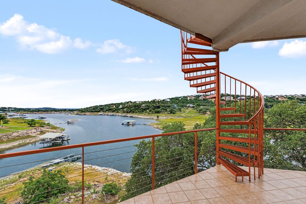 Spiral staircase on a balcony overlooking a scenic river and lush green hills under a partly cloudy sky.