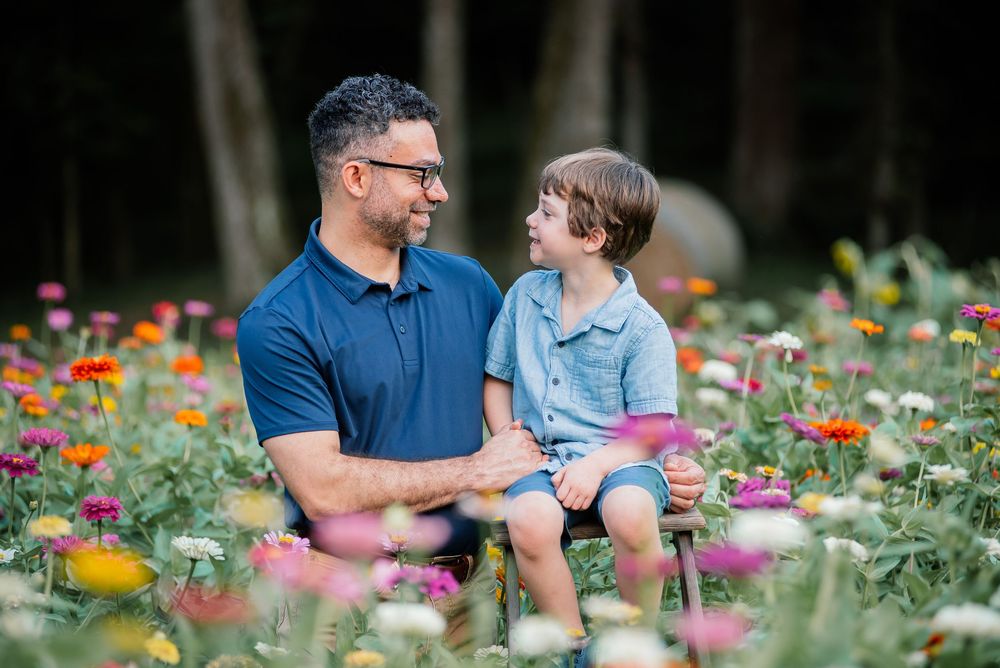 A father holds his young son among colorful zinnia flowers in a garden, both smiling at each other.
