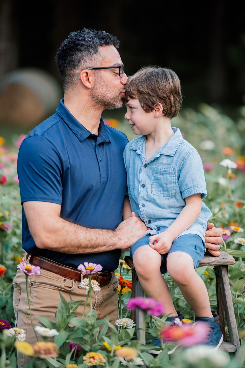 A father kisses his young son on the forehead while sitting together in a colorful flower field.