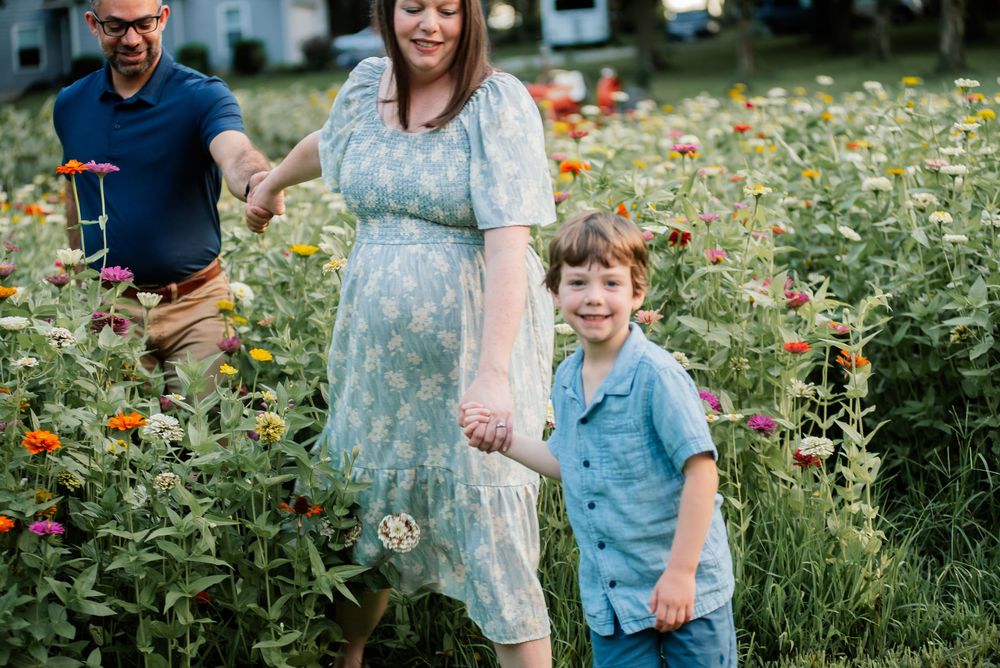 Family of three walks through a colorful flower field, pregnant mother in floral dress holding son's hand outdoors.