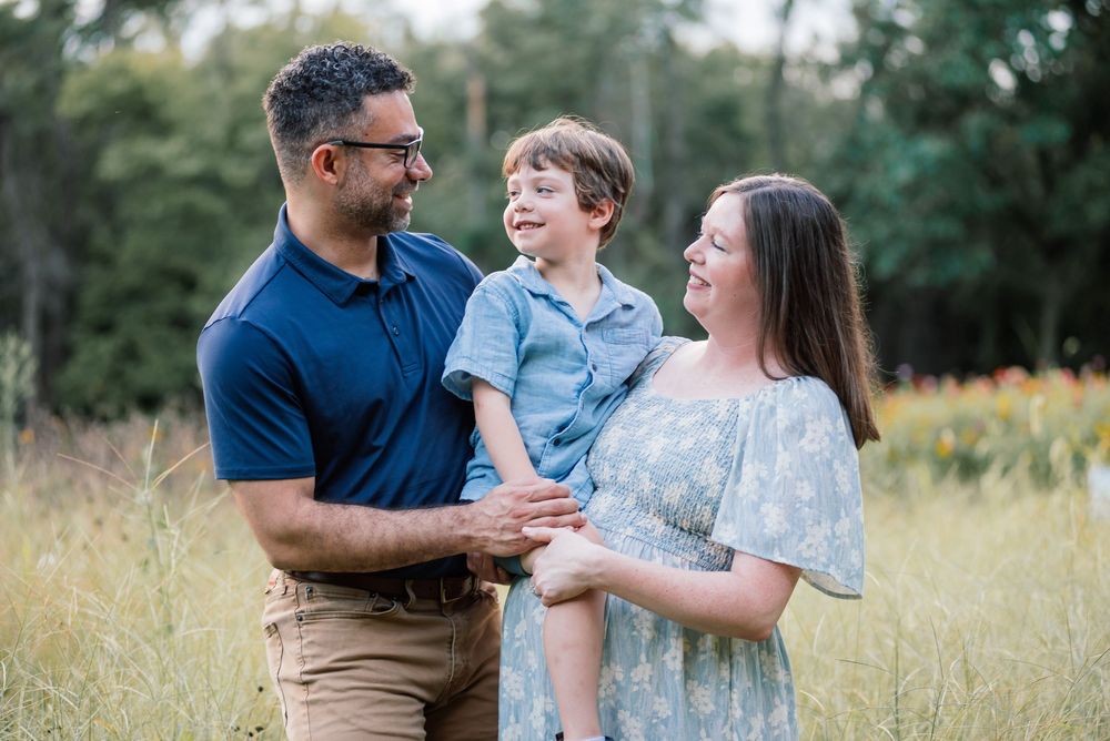Family of three posing outdoors in a golden field, parents smiling at toddler boy, lush green trees in background.