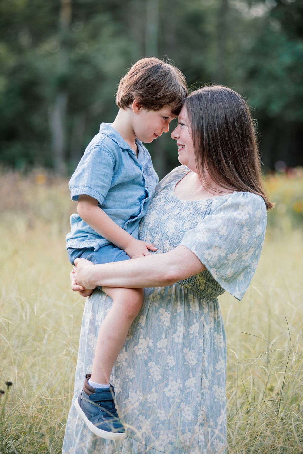 A mother in a floral dress holds her young son in a sunlit field, their foreheads touching tenderly.