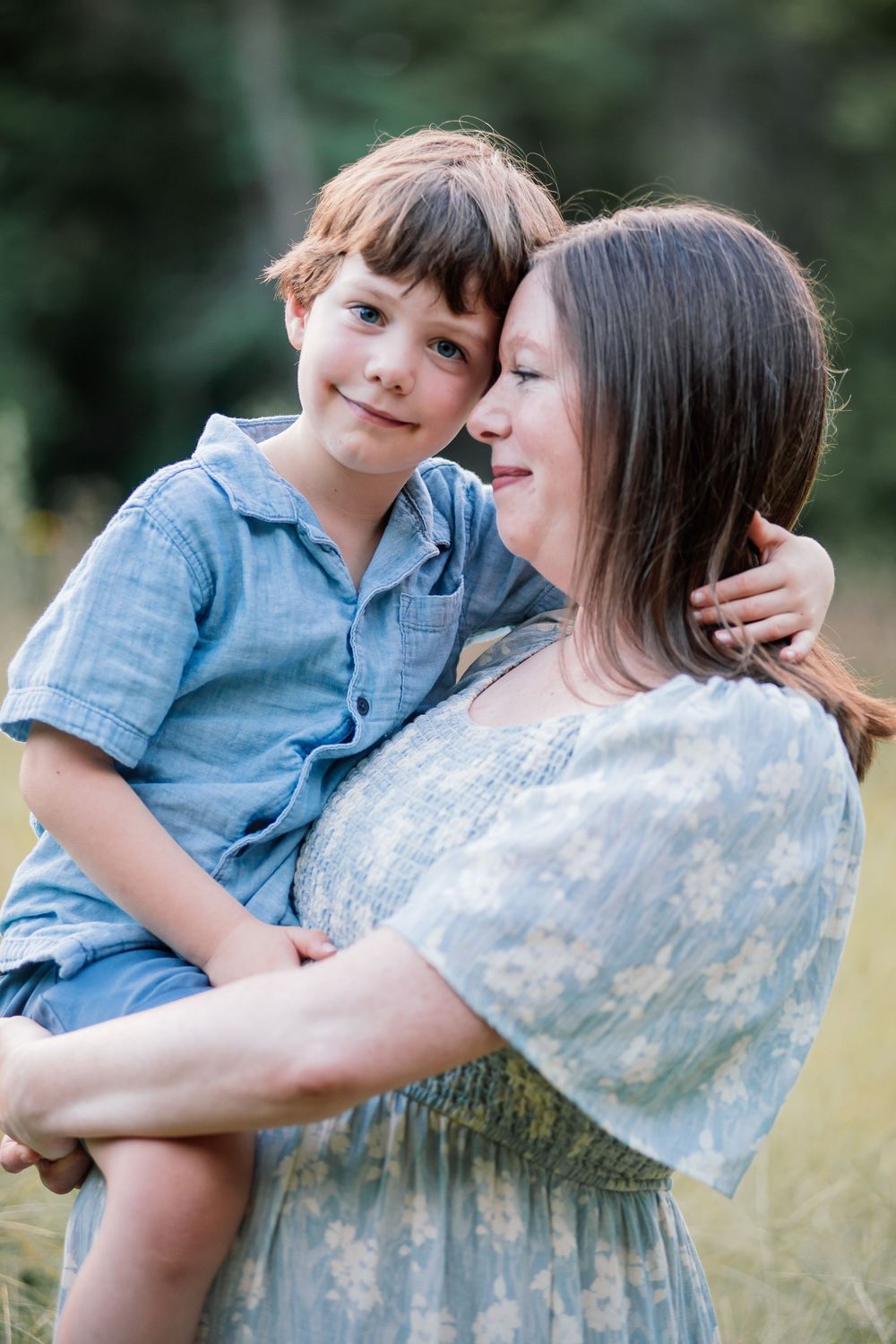A mother in a floral dress holds her smiling young son in a blue outfit outdoors in a green field.