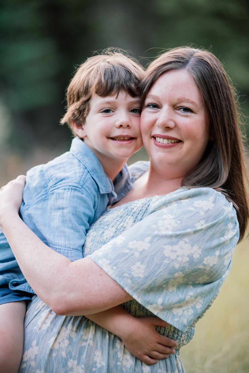 A smiling woman in a floral dress hugs a young boy in a blue shirt outdoors in a sunny field.