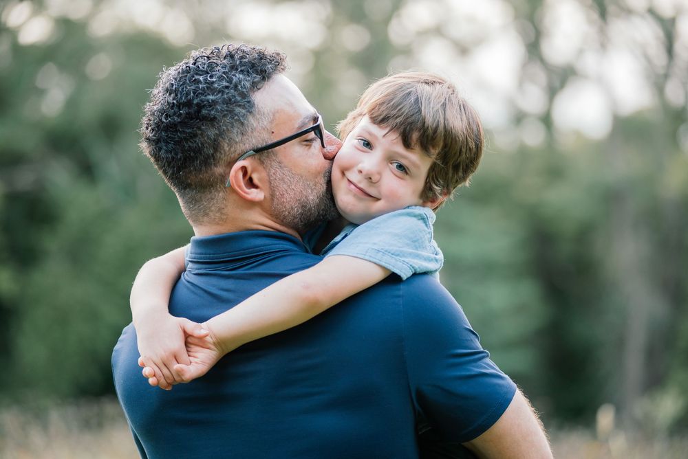 A father holding his smiling young son outdoors, sharing a warm embrace in a green natural setting.