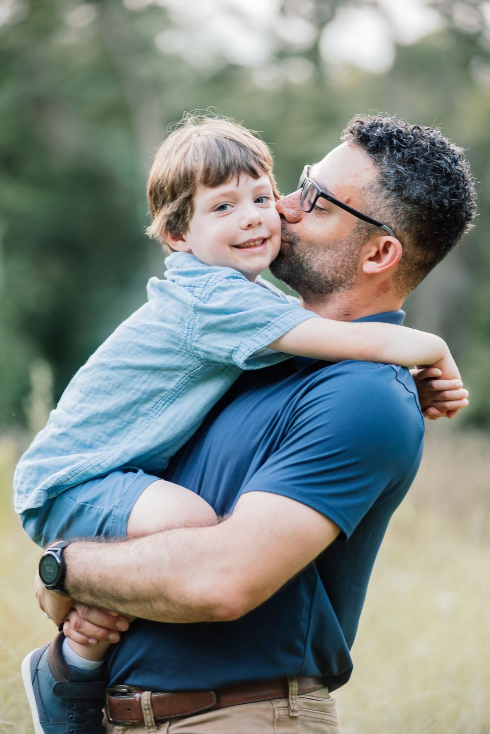 A father kisses his smiling young son on the cheek while holding him outdoors in a green field.