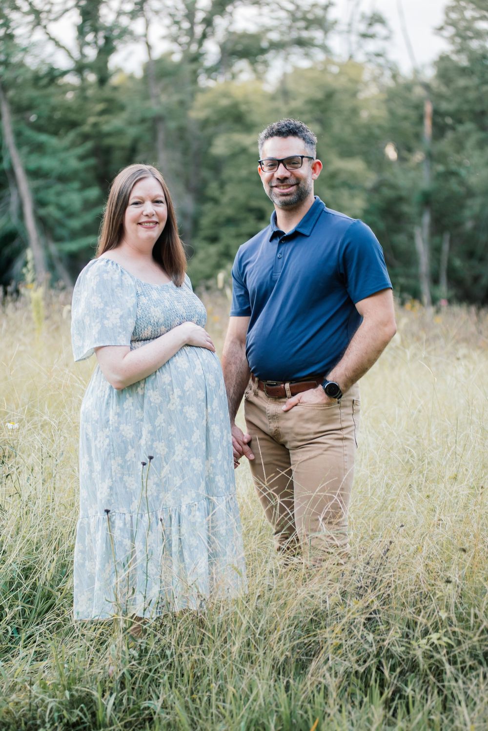 Pregnant woman in floral dress and man in blue polo pose together in a sunny meadow with trees in background.