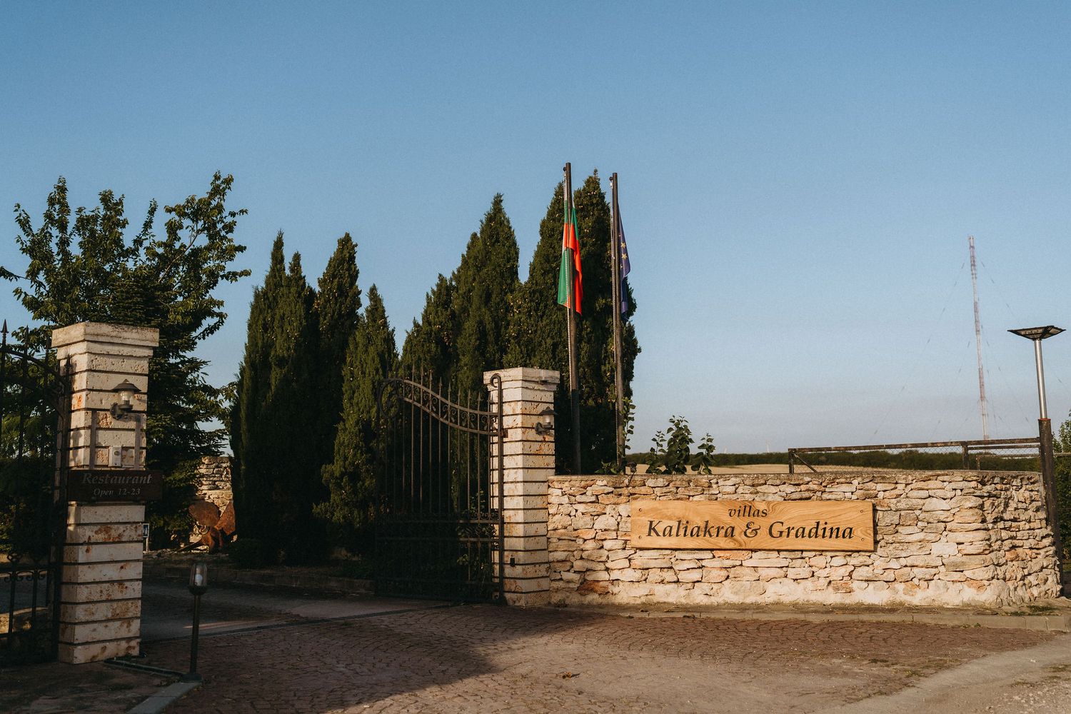 Stone entrance gate of Kallakra & Gradina venue with cypress trees and flag at sunset.