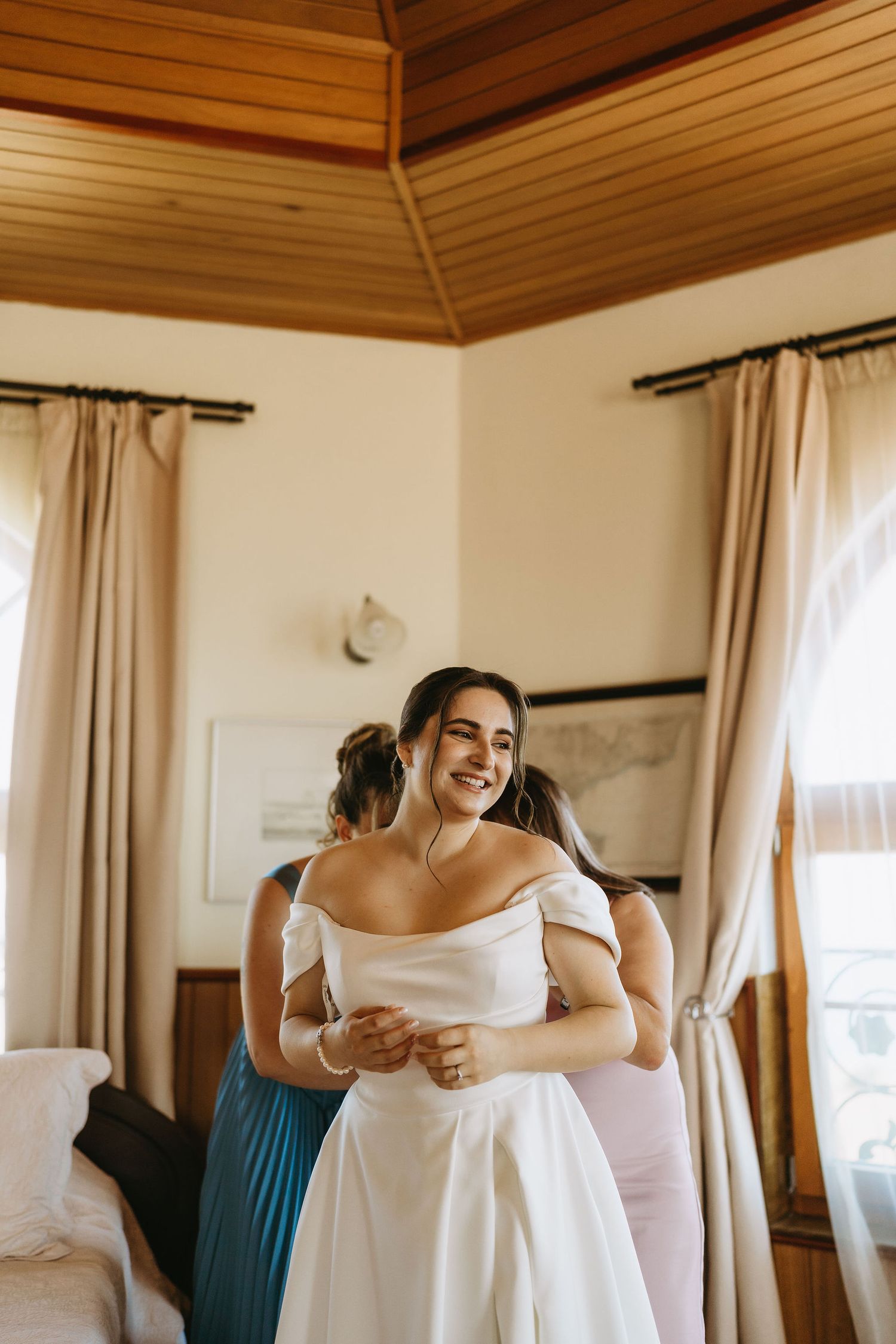 Smiling bride in white off-shoulder gown being helped by bridesmaids in a warmly lit room.