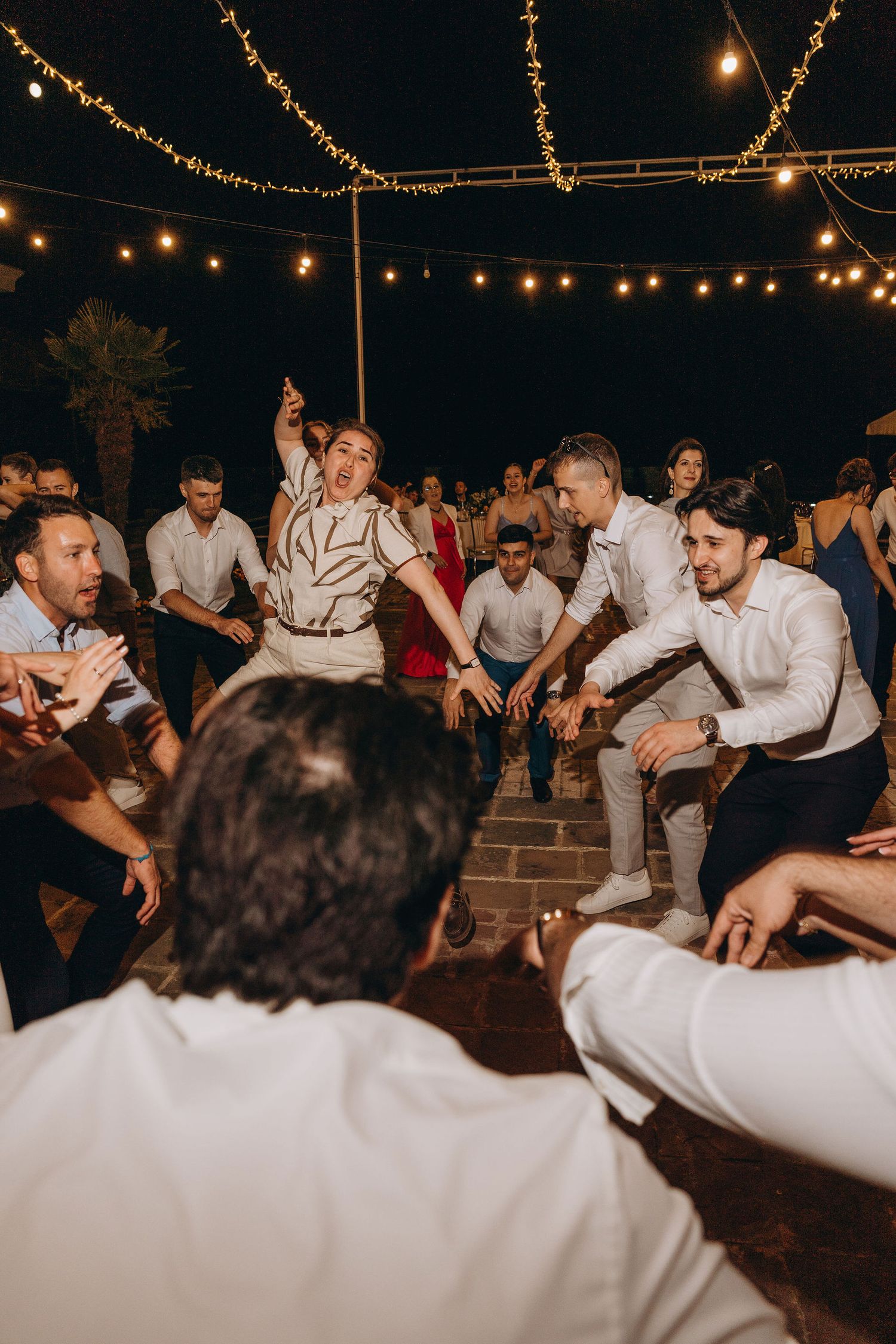 Guests dancing energetically at an outdoor evening wedding reception under warm string lights.