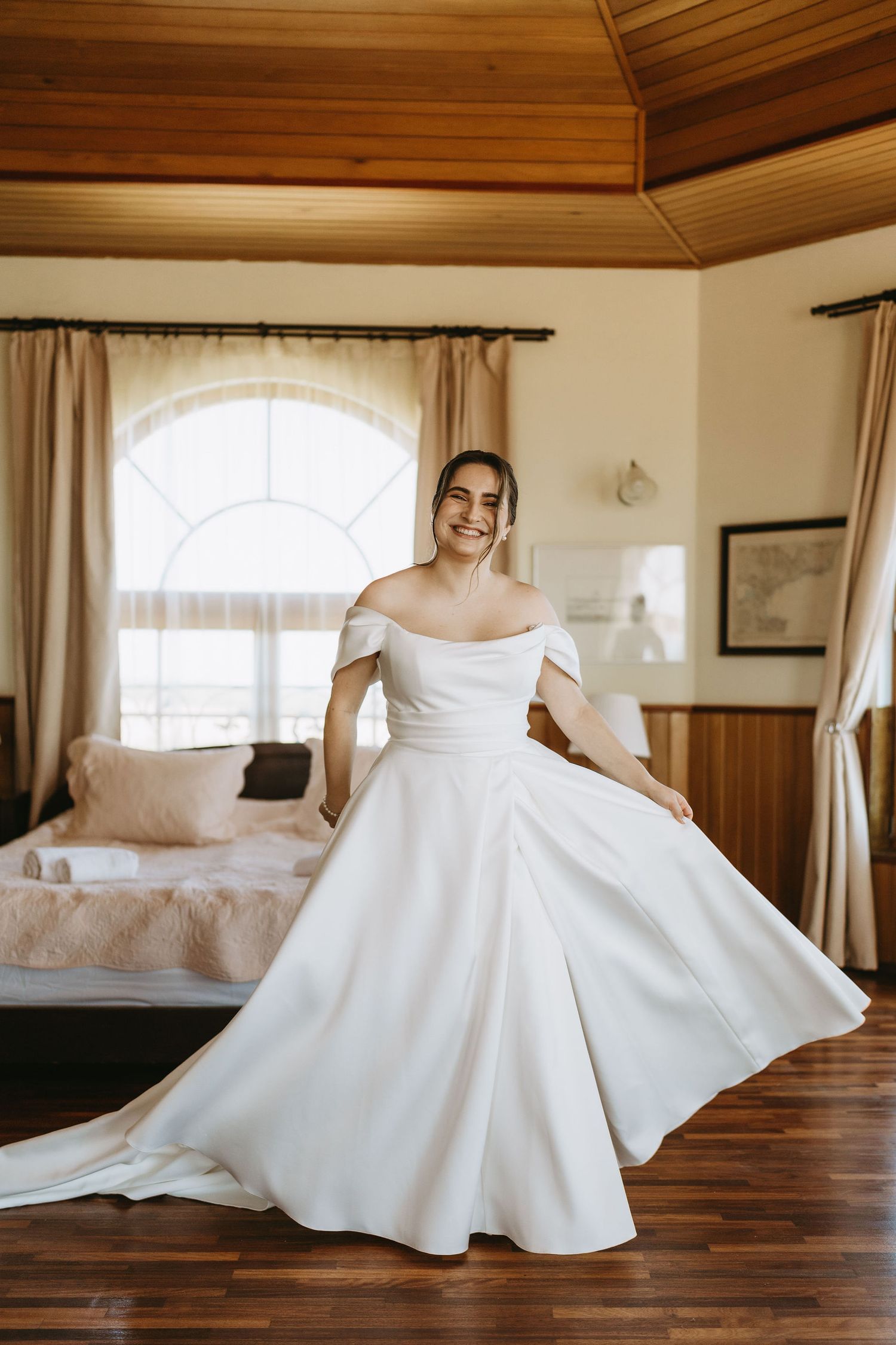 Smiling bride in off-shoulder white ball gown twirling in elegant wood-paneled room with arched window.