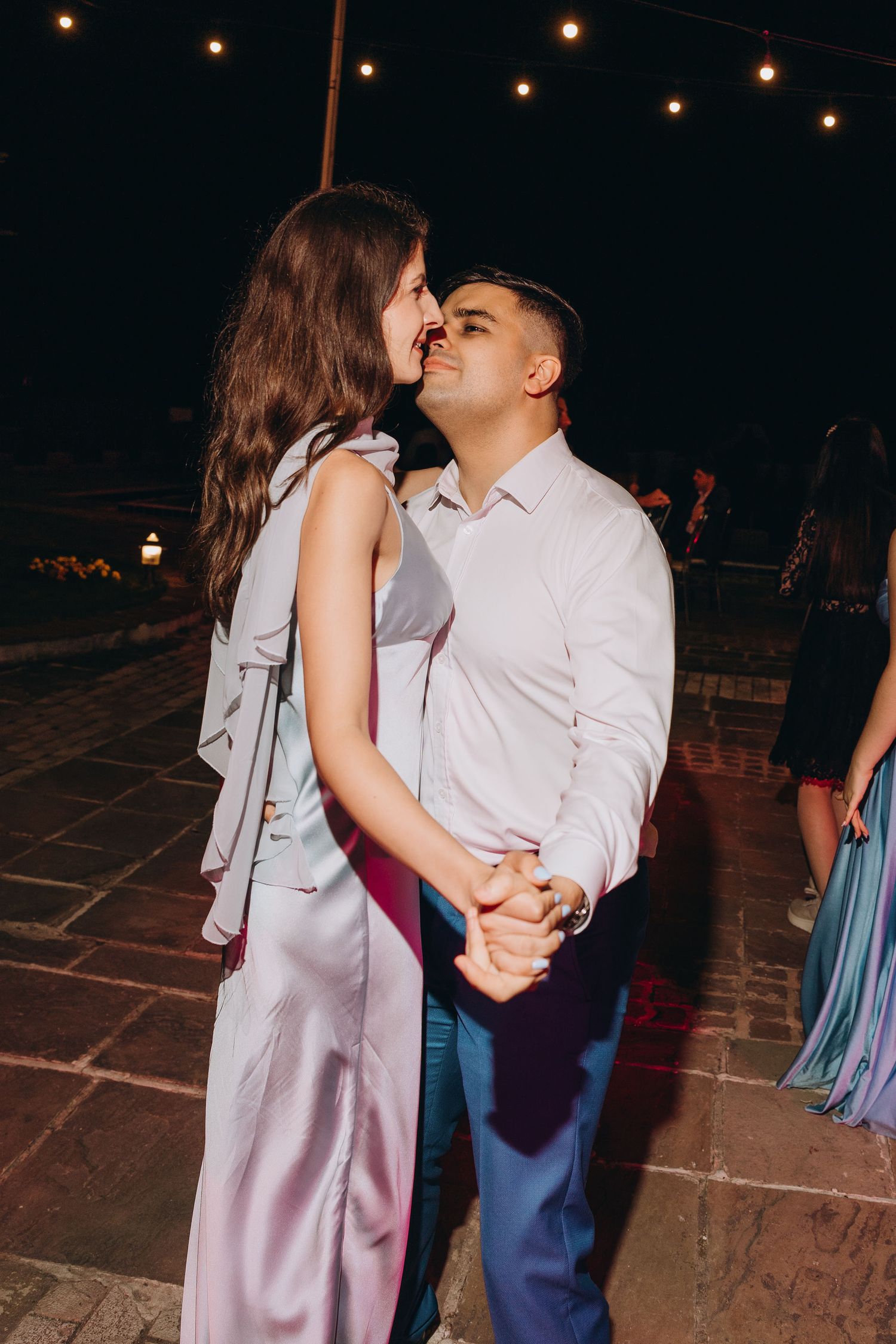 Couple dancing closely at an outdoor evening event, string lights glowing above them.