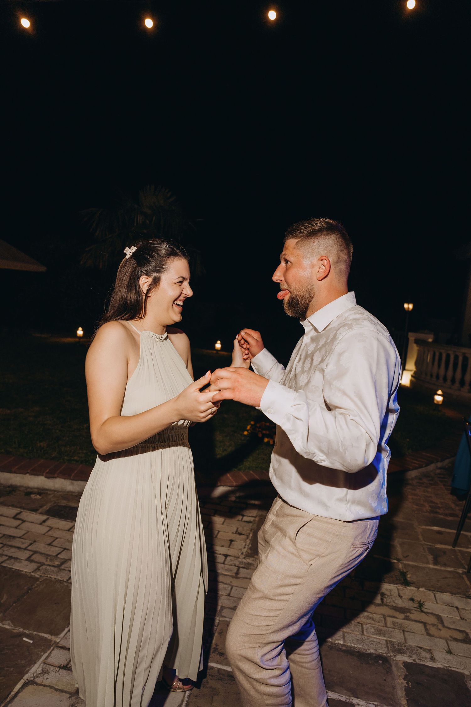 Couple dancing joyfully outdoors at night, string lights glowing, woman in sage dress, man in white shirt.