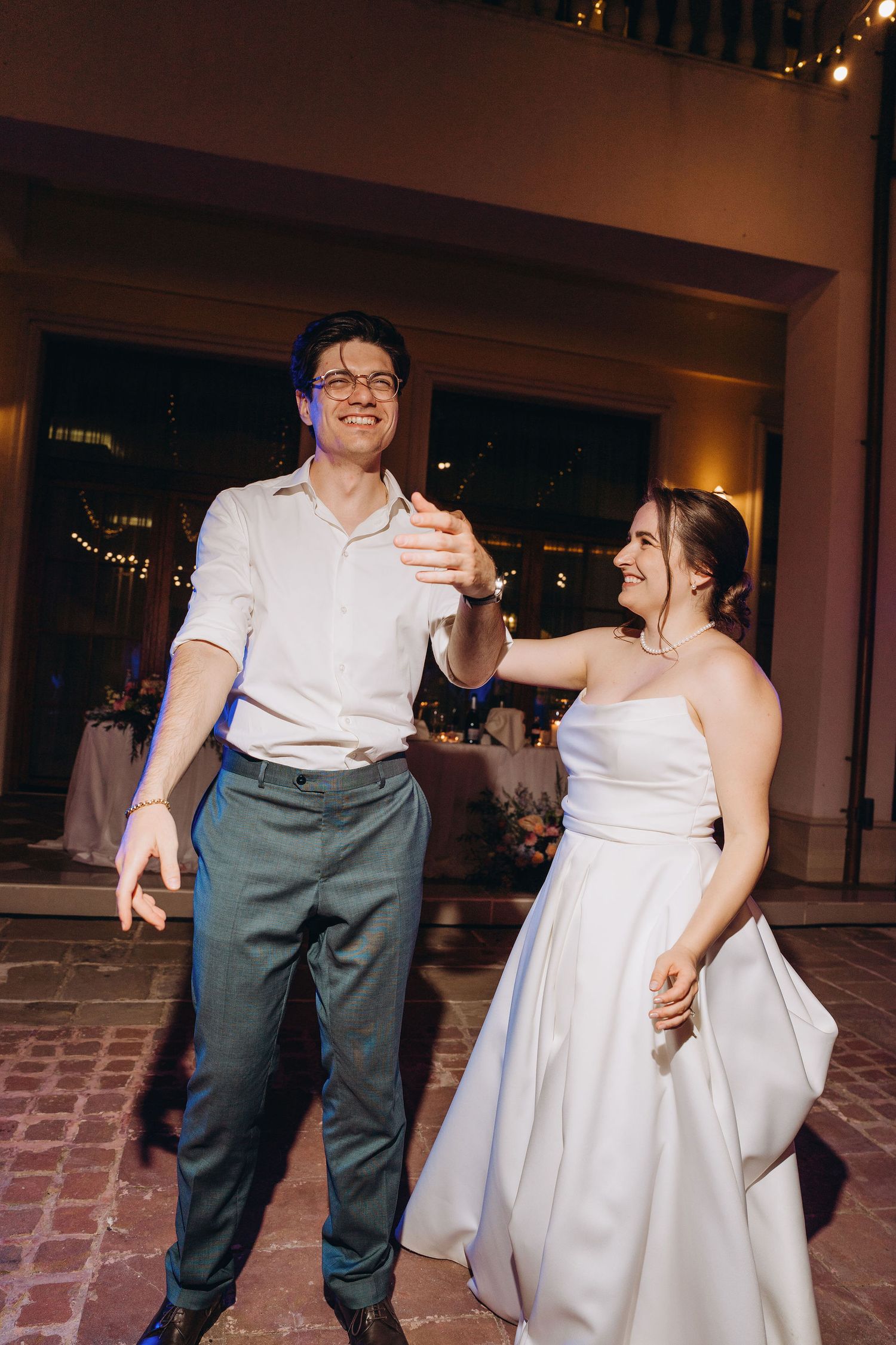 A joyful bride in a white strapless gown and groom in white shirt dance together at their wedding reception.