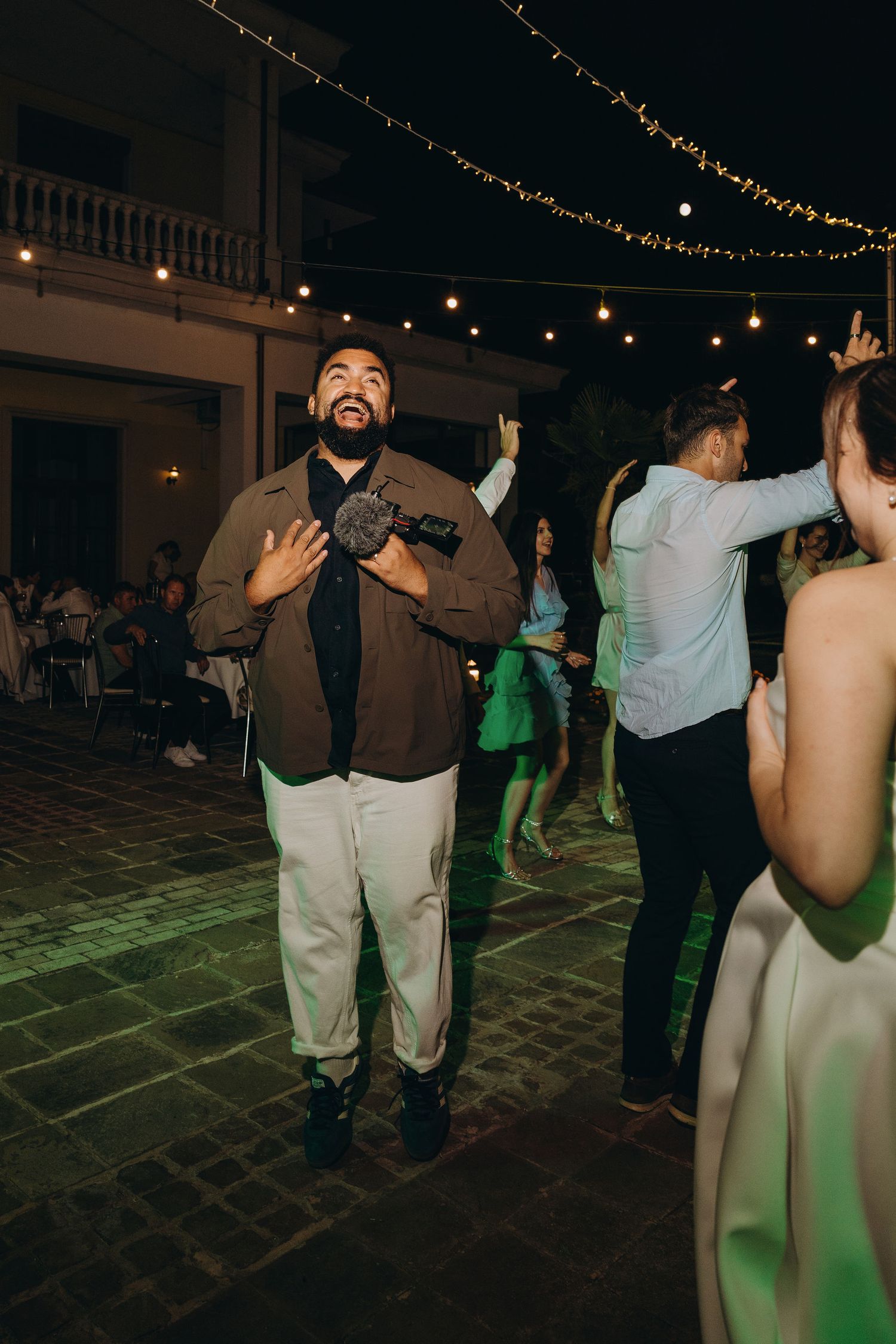 Man smiling and dancing at an outdoor evening wedding reception with string lights and guests.