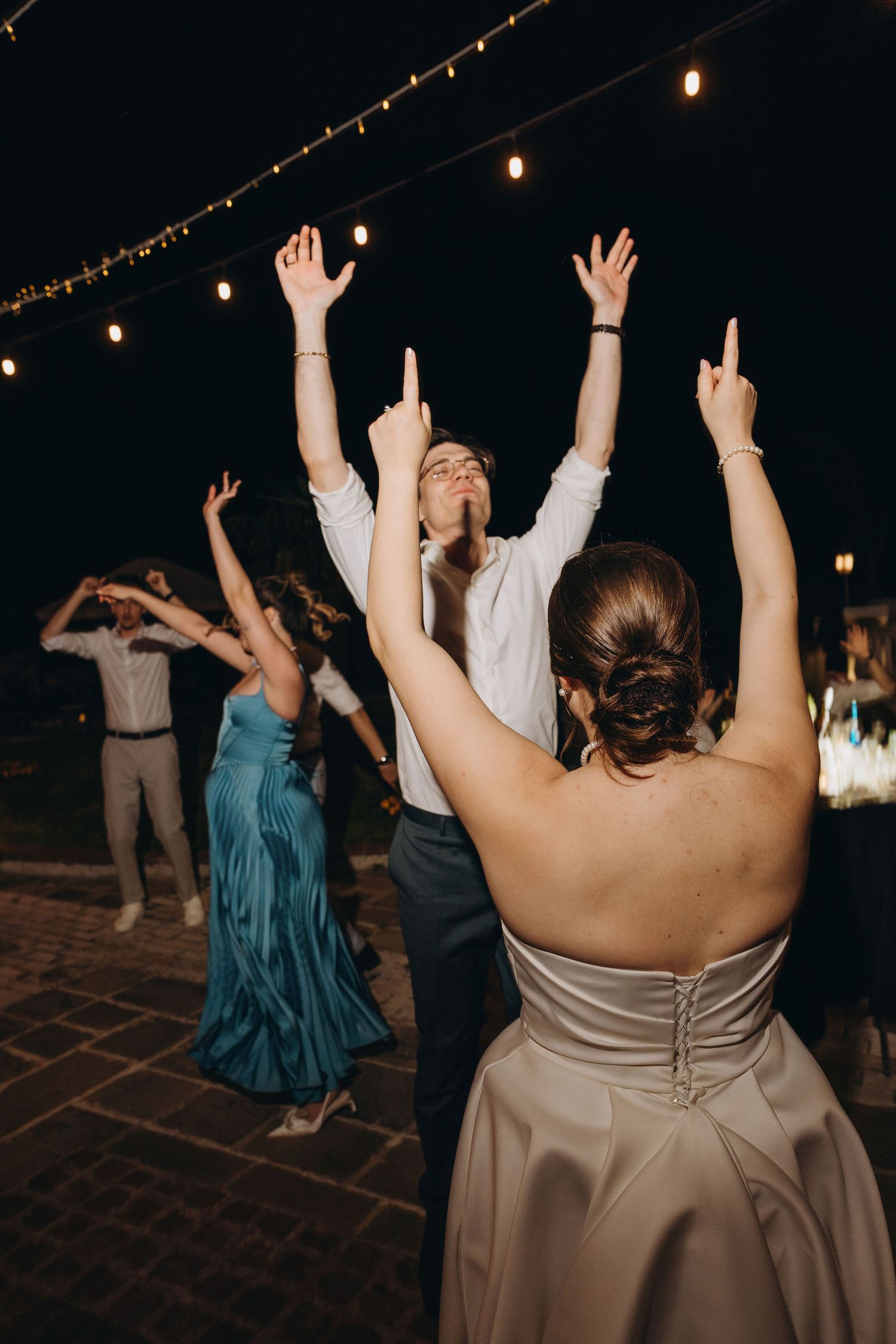 Guests dancing outdoors at a wedding reception under string lights, wearing formal attire including a satin gown.