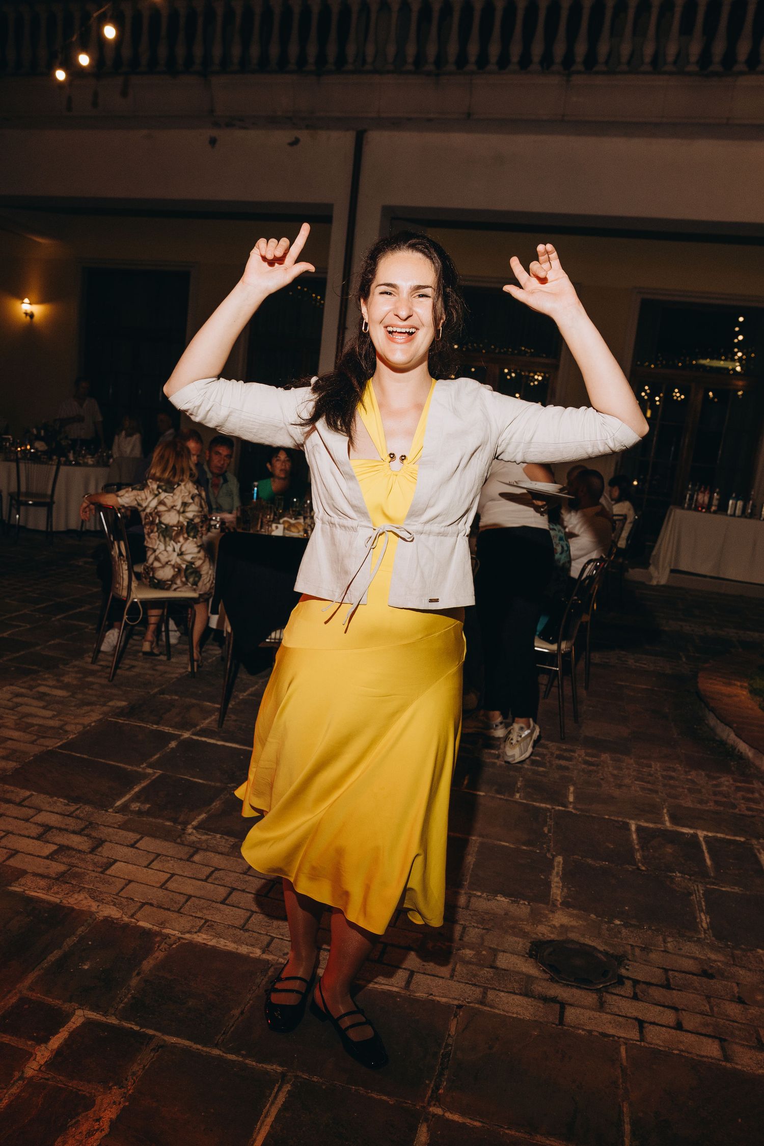 Woman in yellow skirt and white jacket dancing joyfully at an indoor evening event or wedding reception.