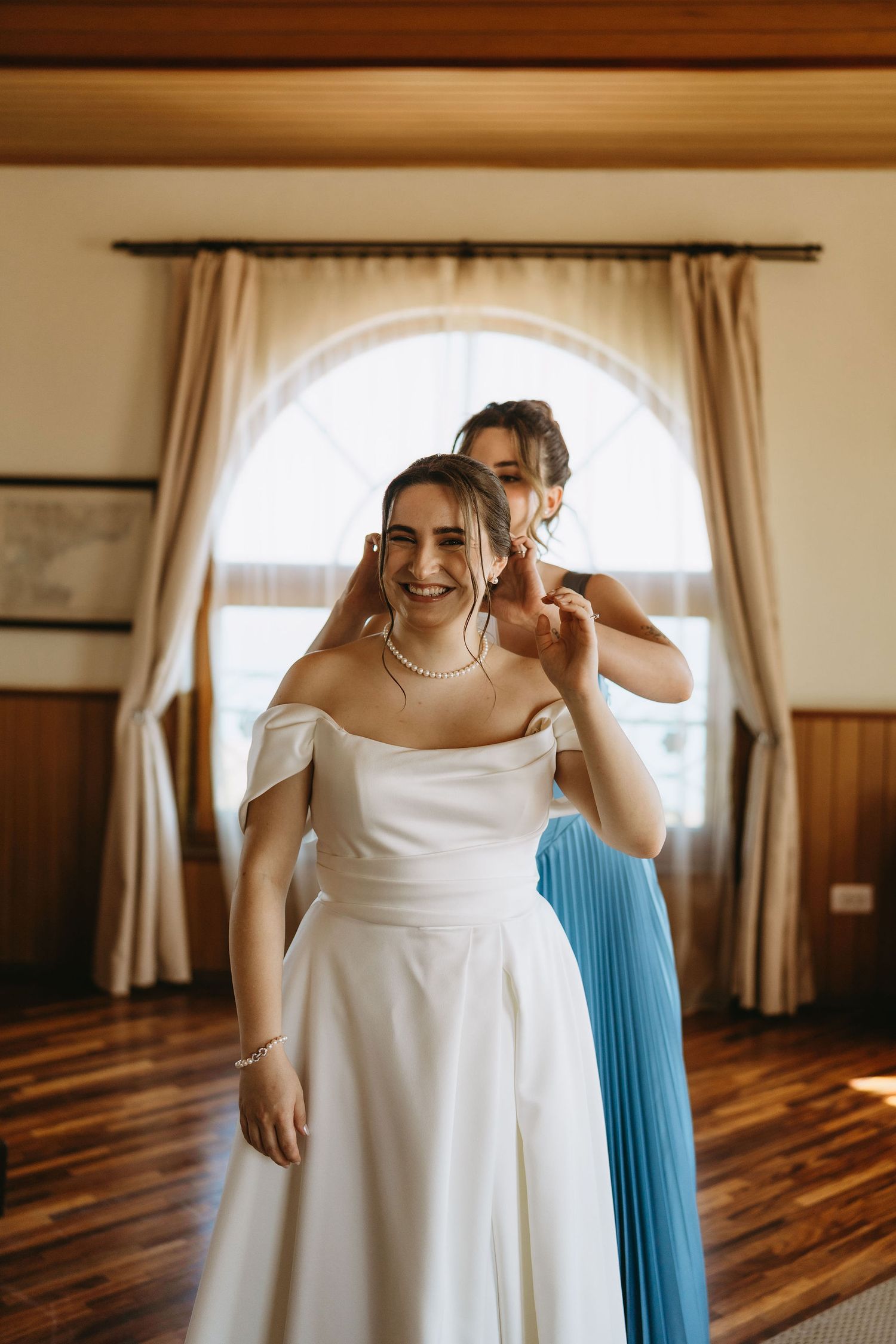 Bridesmaid in blue dress helping bride in white off-shoulder gown with jewelry in elegant wood-paneled room.