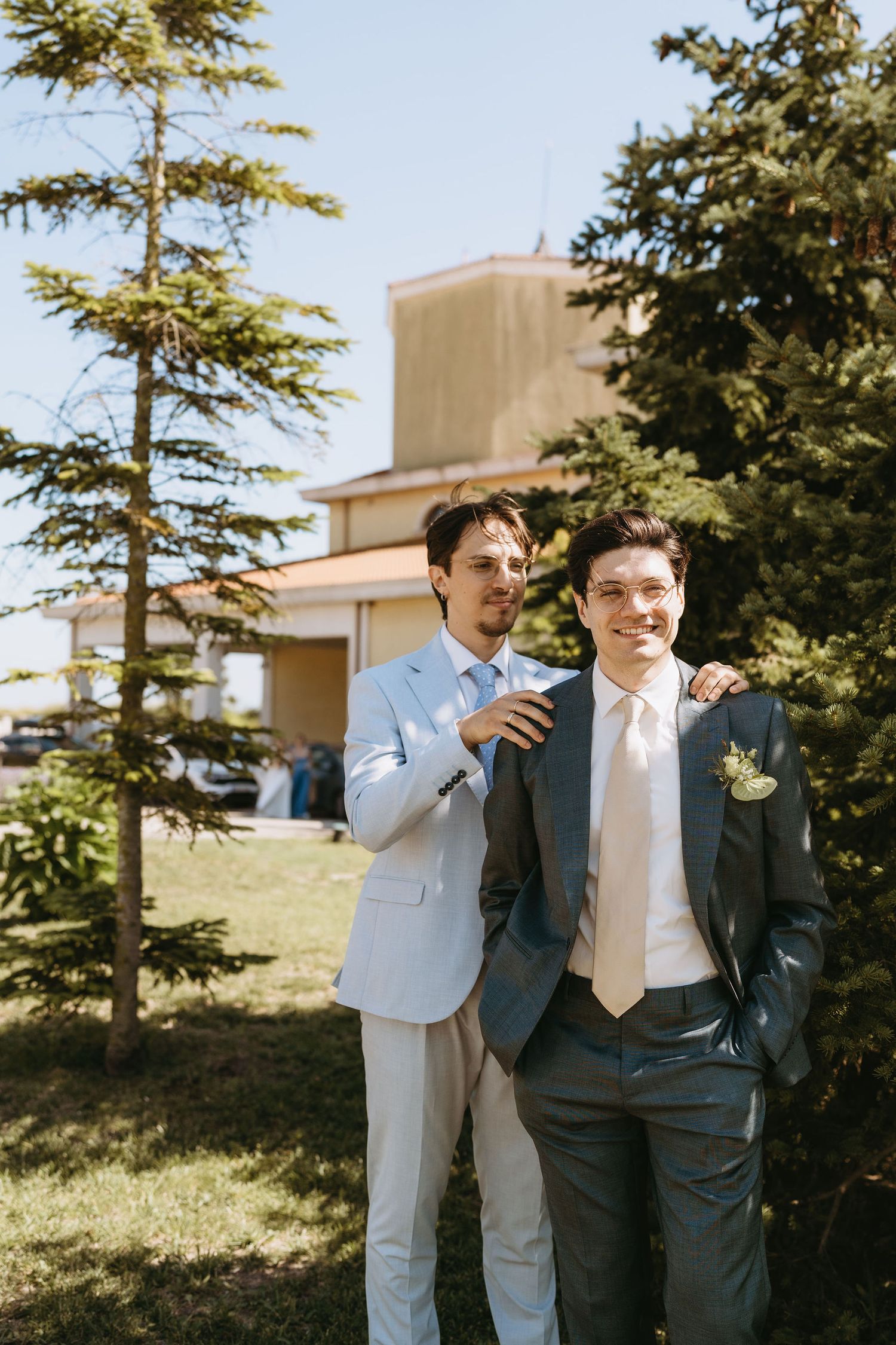 Two grooms in suits stand outdoors near evergreen trees on their wedding day, one wearing a boutonniere.