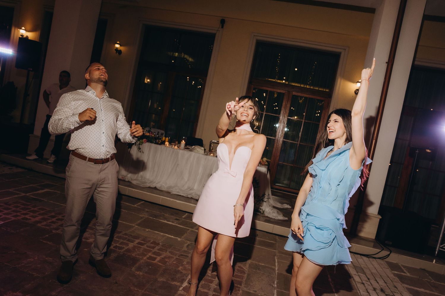 Three guests dancing joyfully at an indoor wedding reception, woman in white dress and floral crown centerpiece.
