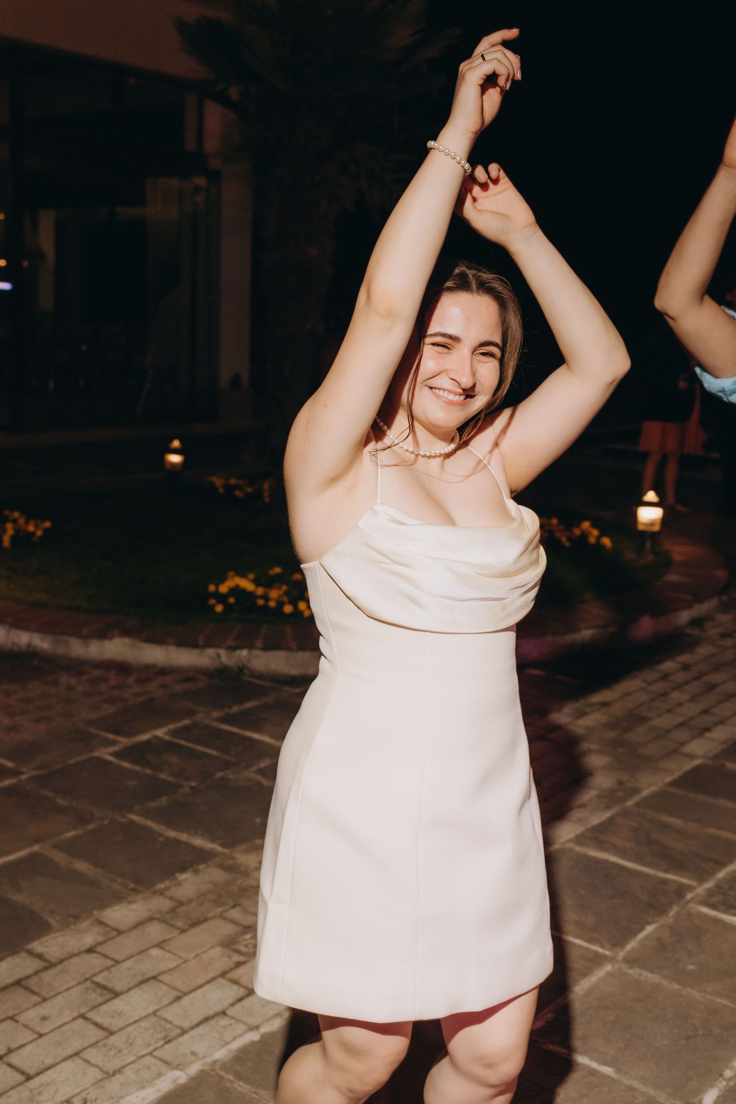 Young woman in white mini dress dancing with arms raised at an outdoor evening event.