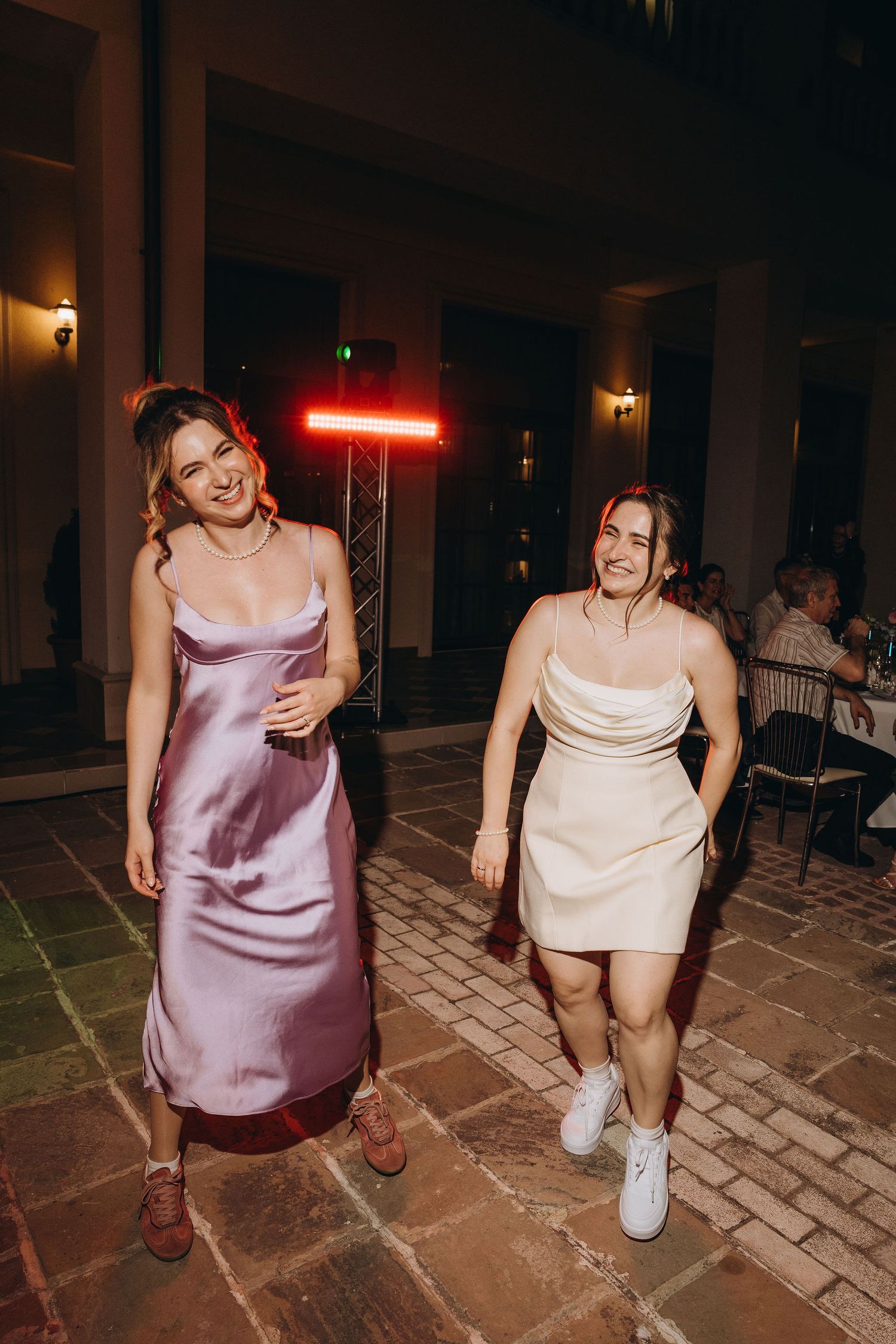 Two women in satin slip dresses smiling and walking at an evening event on a cobblestone patio.