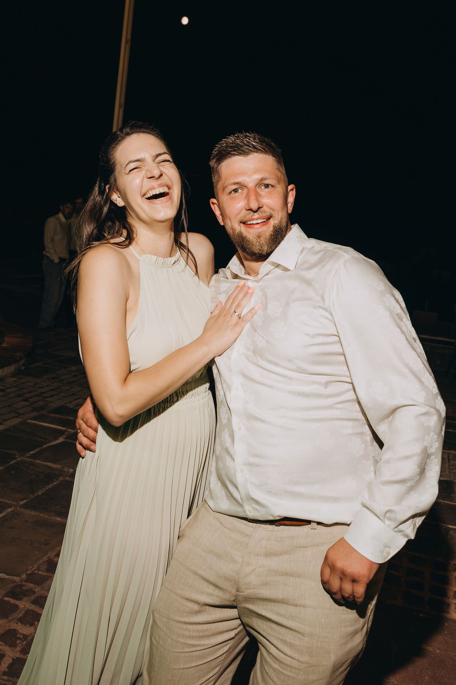 A laughing couple in elegant attire poses together at an outdoor evening wedding reception.