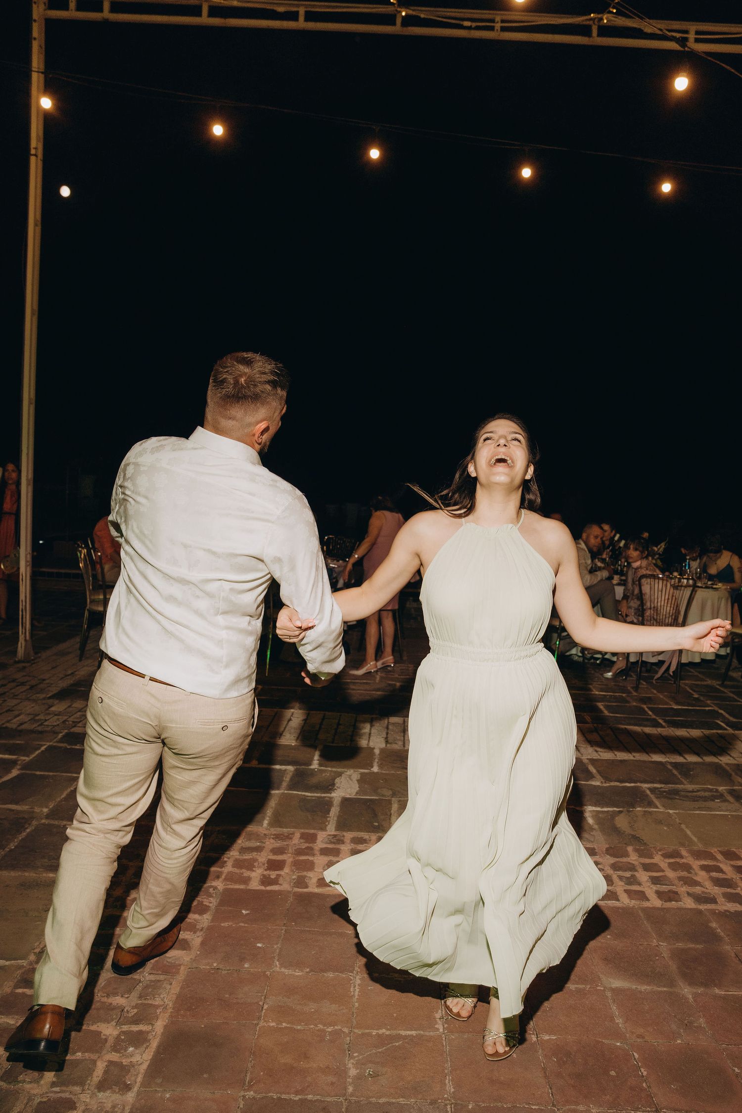 Bride in white halter gown laughs joyfully while dancing with groom in khaki suit under string lights at outdoor wedding reception.