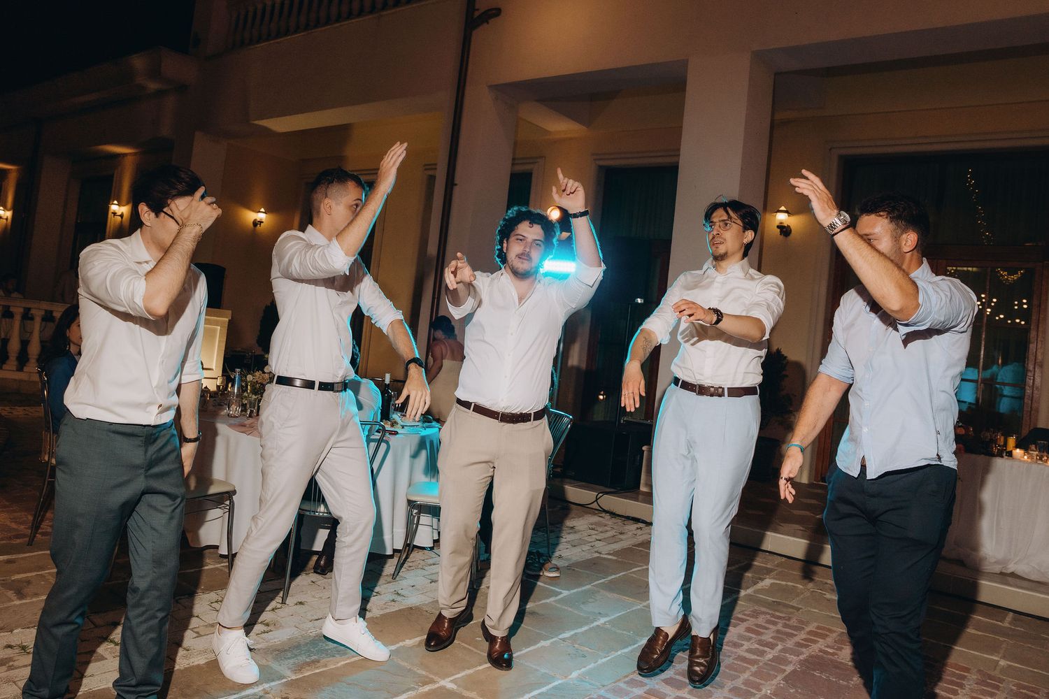 Groomsmen dancing together outdoors at a wedding reception at night, wearing white dress shirts and khaki pants.