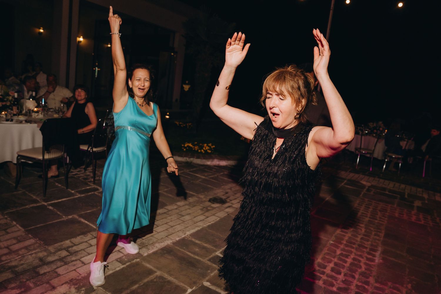 Two women dancing joyfully with arms raised at an outdoor evening wedding reception celebration.