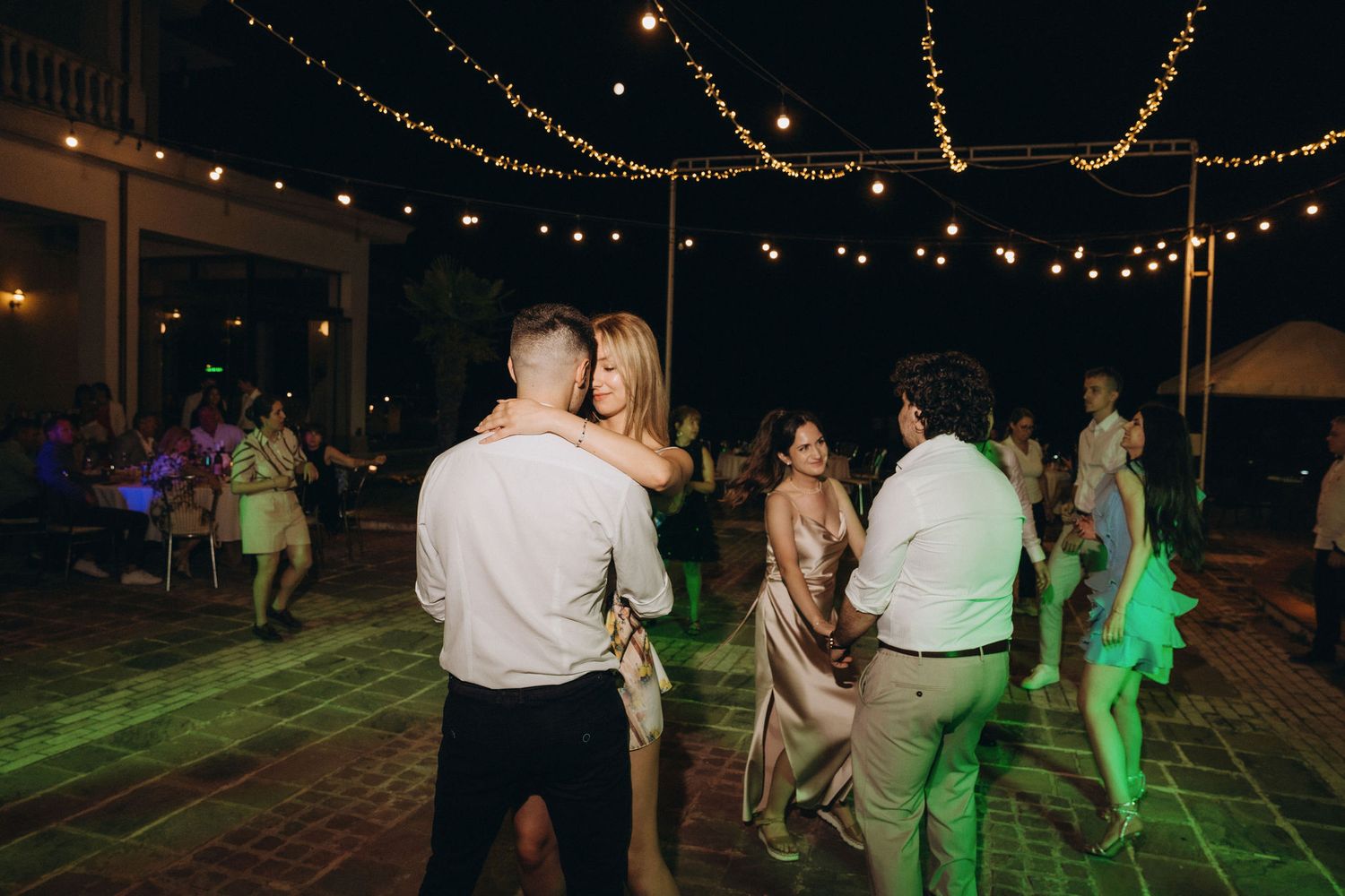Couples dancing outdoors at a wedding reception under string lights on a green-lit patio at night.