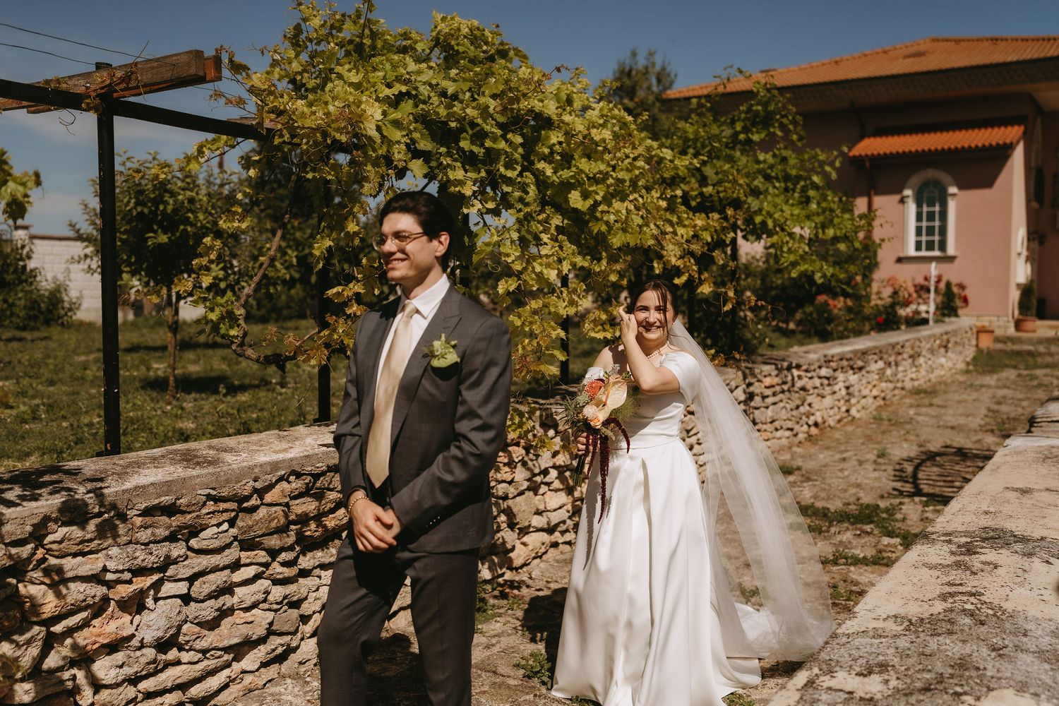 Bride in white gown and groom in gray suit walk outdoors near stone wall and green vineyard at rustic wedding venue.