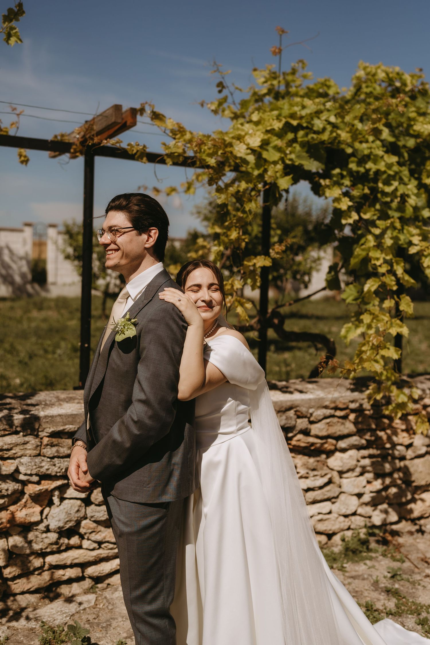 Bride in white gown embraces groom in gray suit outdoors near stone wall and vineyard on sunny wedding day.