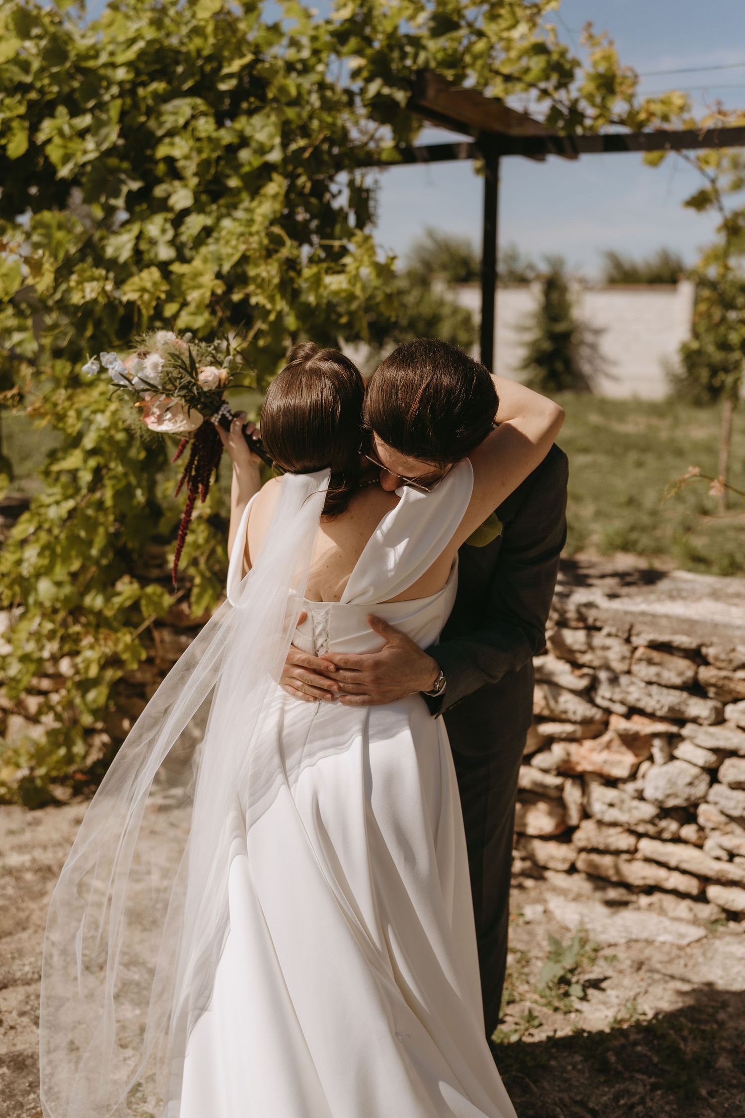 Bride and groom sharing a romantic embrace outdoors, surrounded by lush greenery and rustic stone walls on their wedding day.
