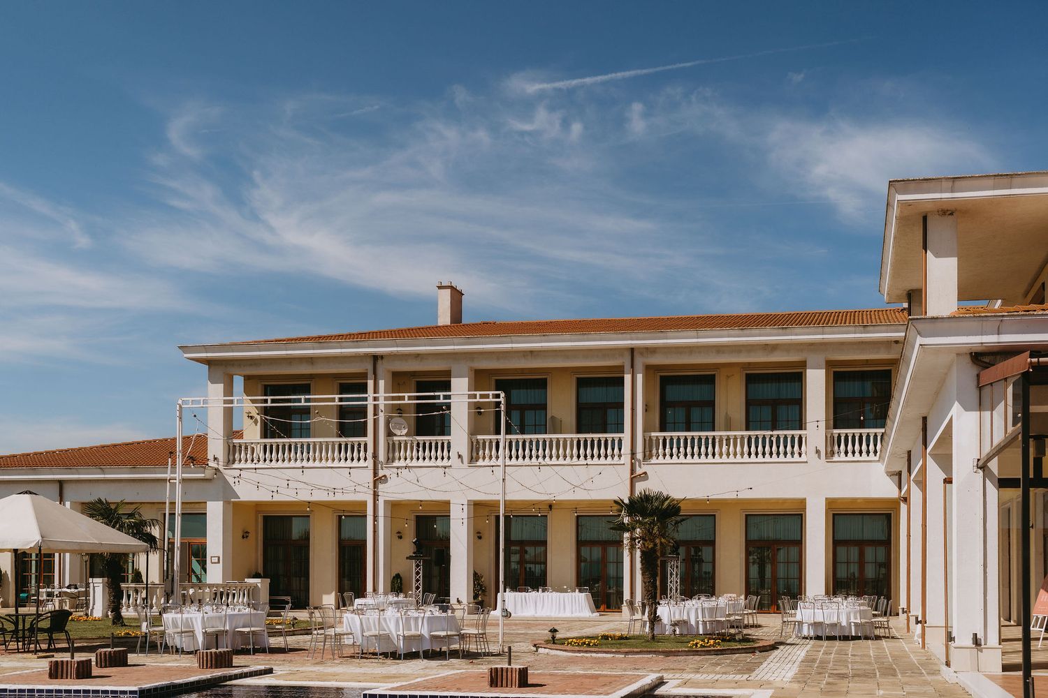 Elegant two-story venue with white balustrade balcony, outdoor seating, and manicured grounds under a blue sky.
