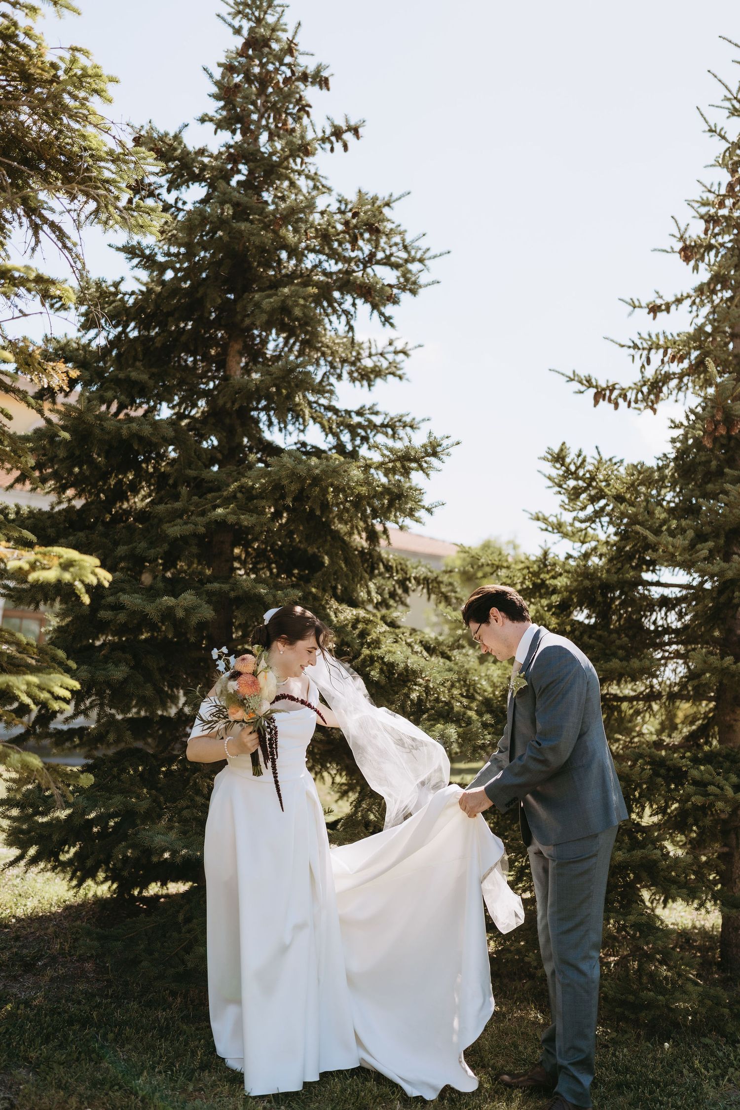 Bride and groom stand outdoors among tall evergreen trees, groom adjusting brides white wedding gown on sunny day.
