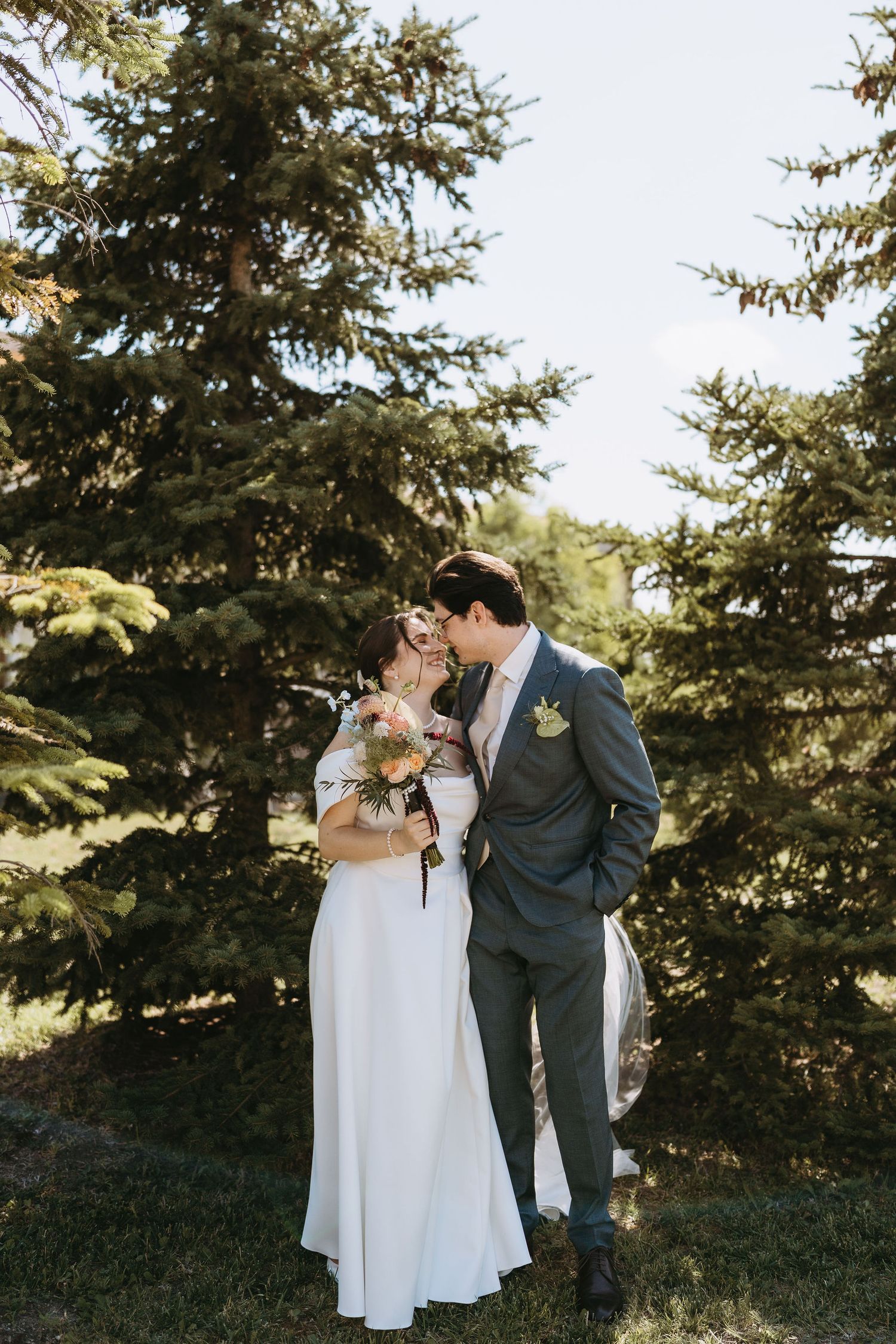 A bride and groom share a romantic kiss outdoors near tall evergreen trees on their wedding day.