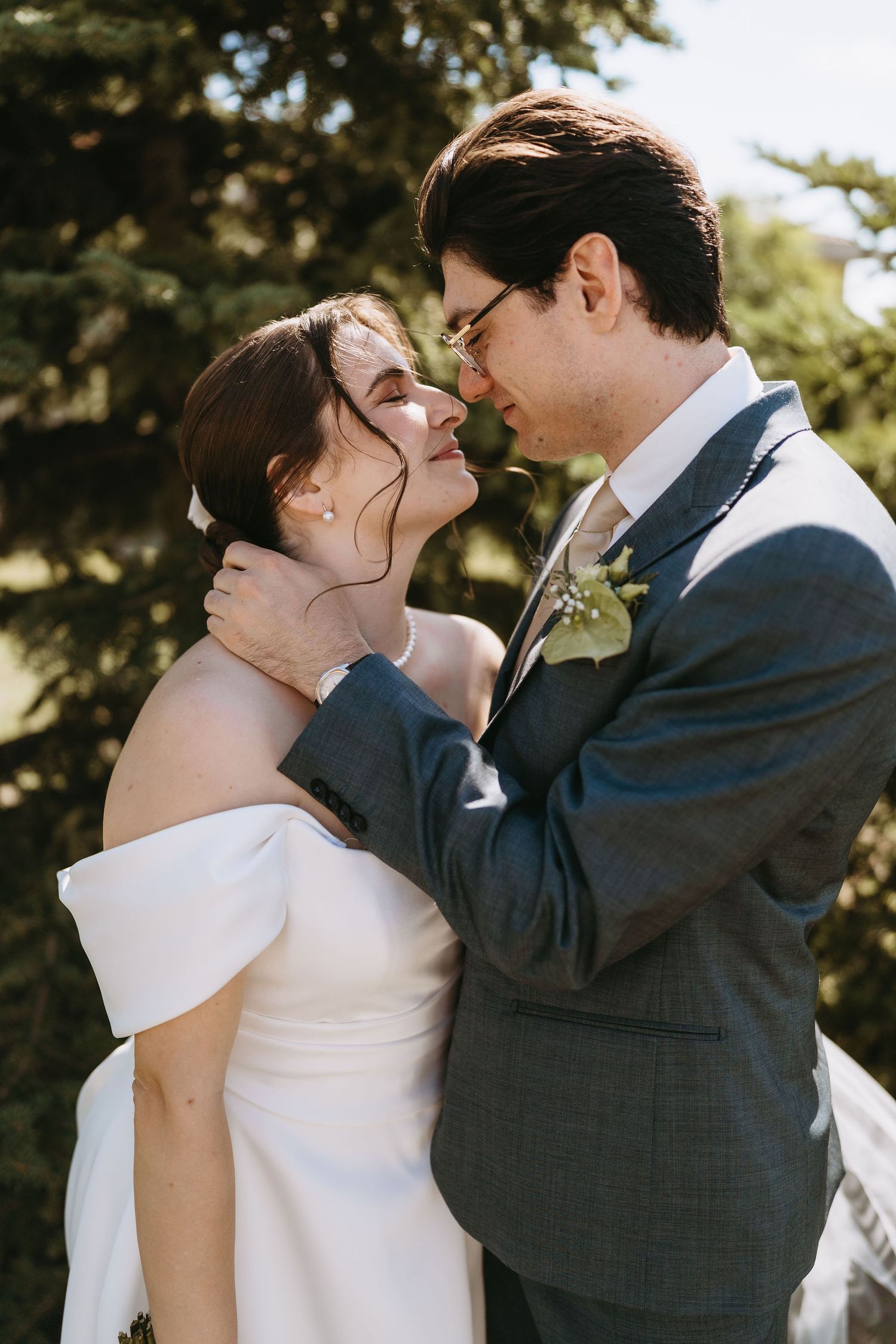 Bride and groom share a romantic moment, touching foreheads outdoors with evergreen trees in the background.