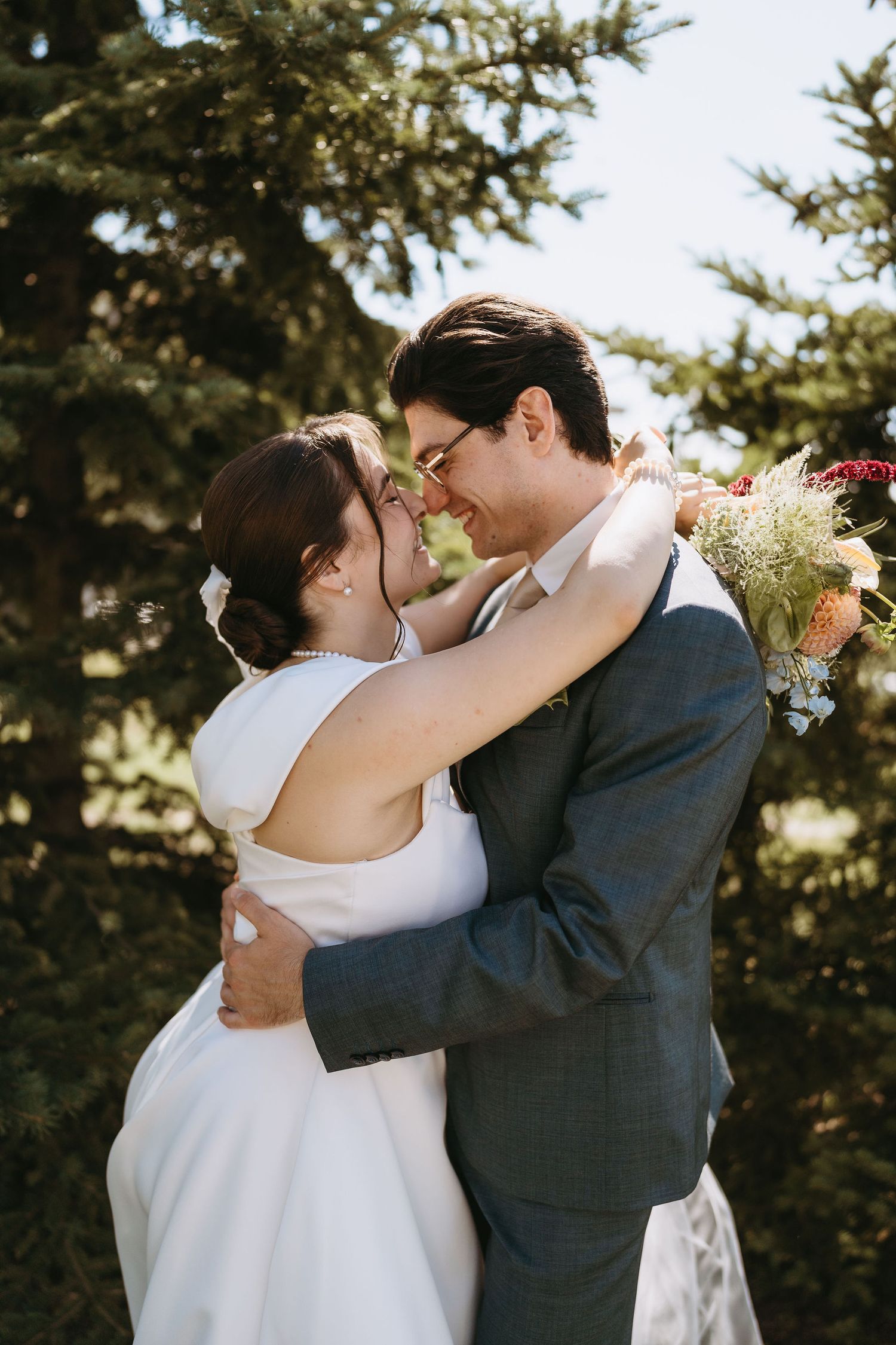 Newlyweds embrace and touch foreheads while bride holds a colorful bouquet, surrounded by lush evergreen trees.