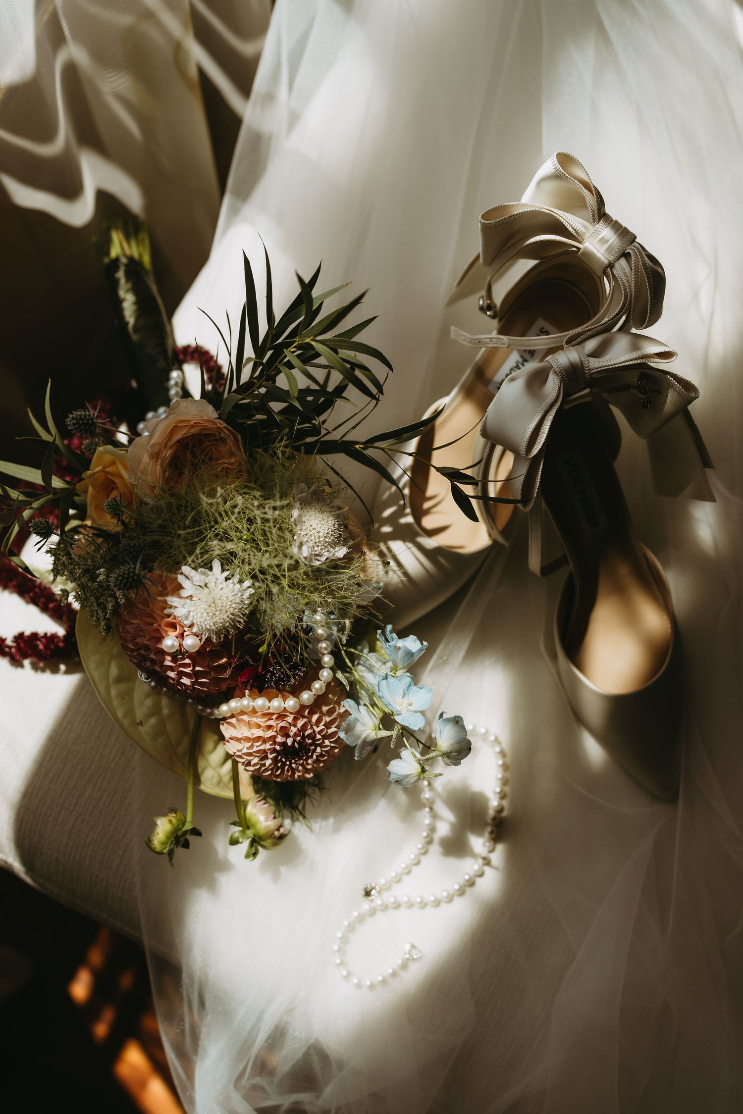 Bridal flat lay with a wildflower bouquet, pearl necklace, and satin heels on white tulle fabric.